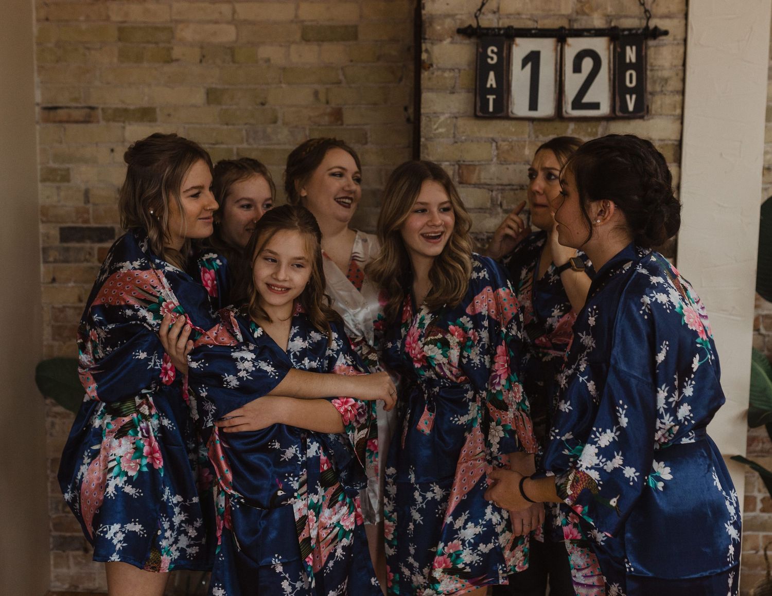 Bridesmaids in matching navy floral robes pose together in a rustic brick-walled room.