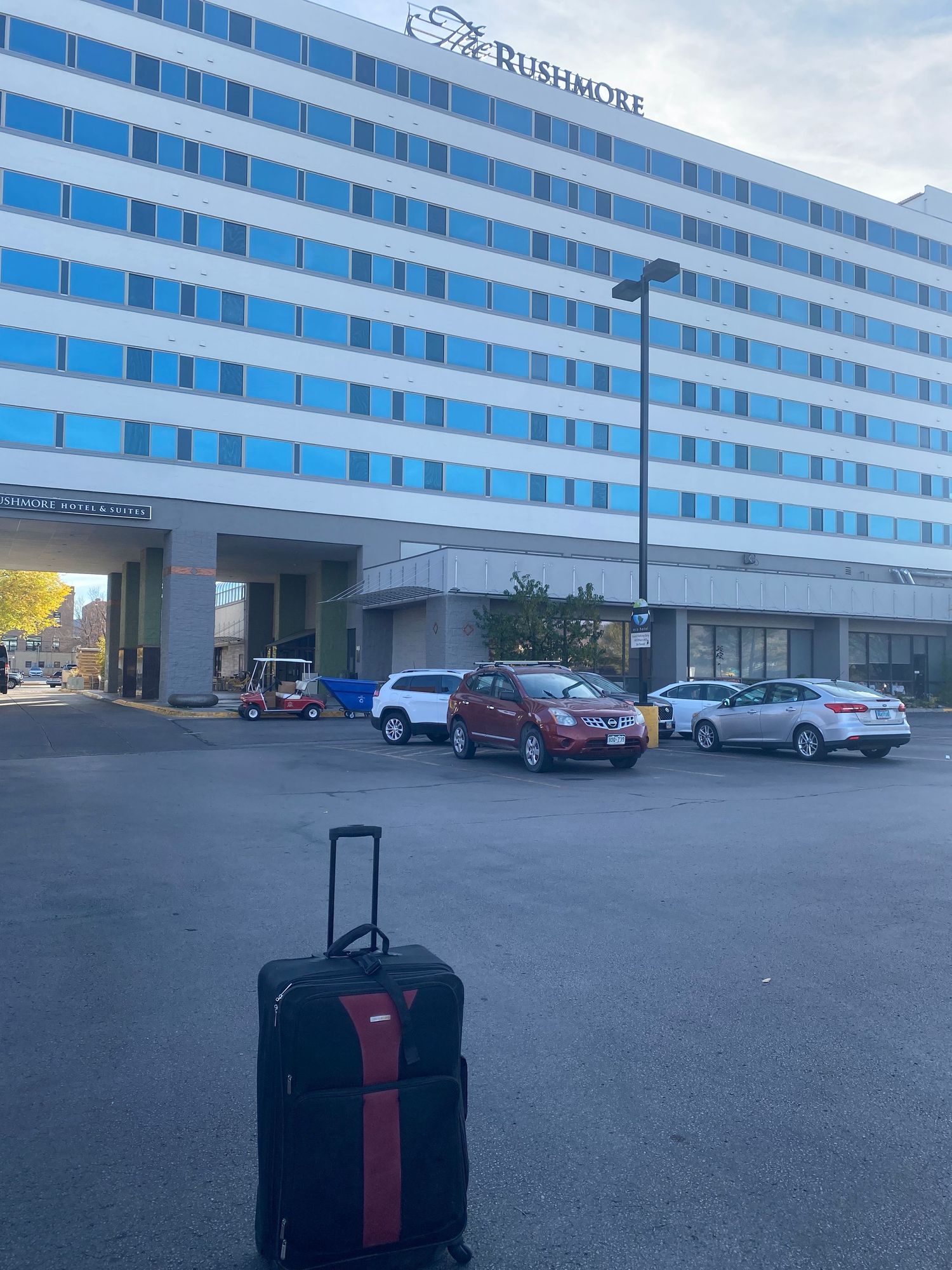 Black suitcase standing in front of a modern Rushmore hotel with parked cars.
