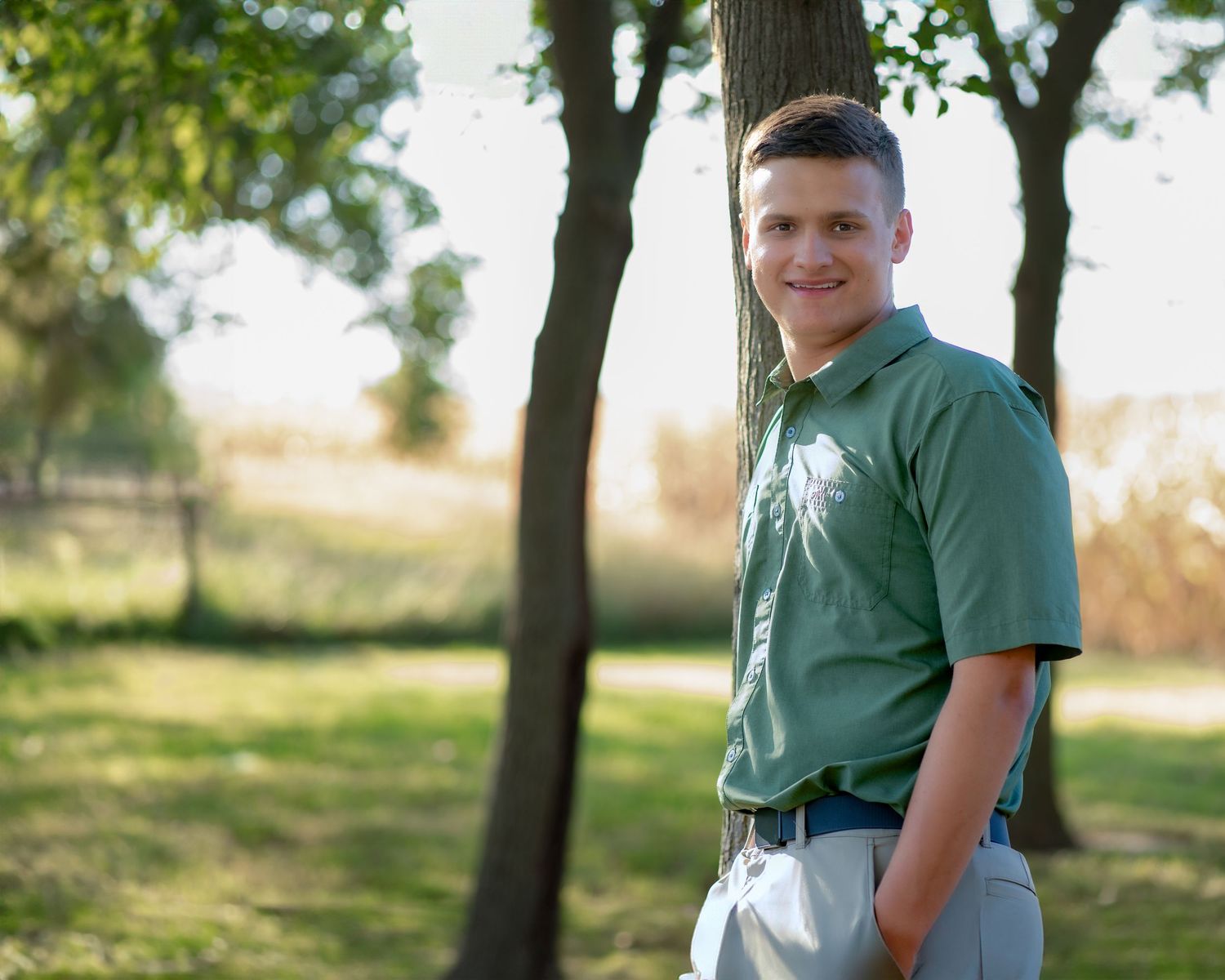 A young individual in a green polo shirt poses outdoors against a sunny, tree-lined background.