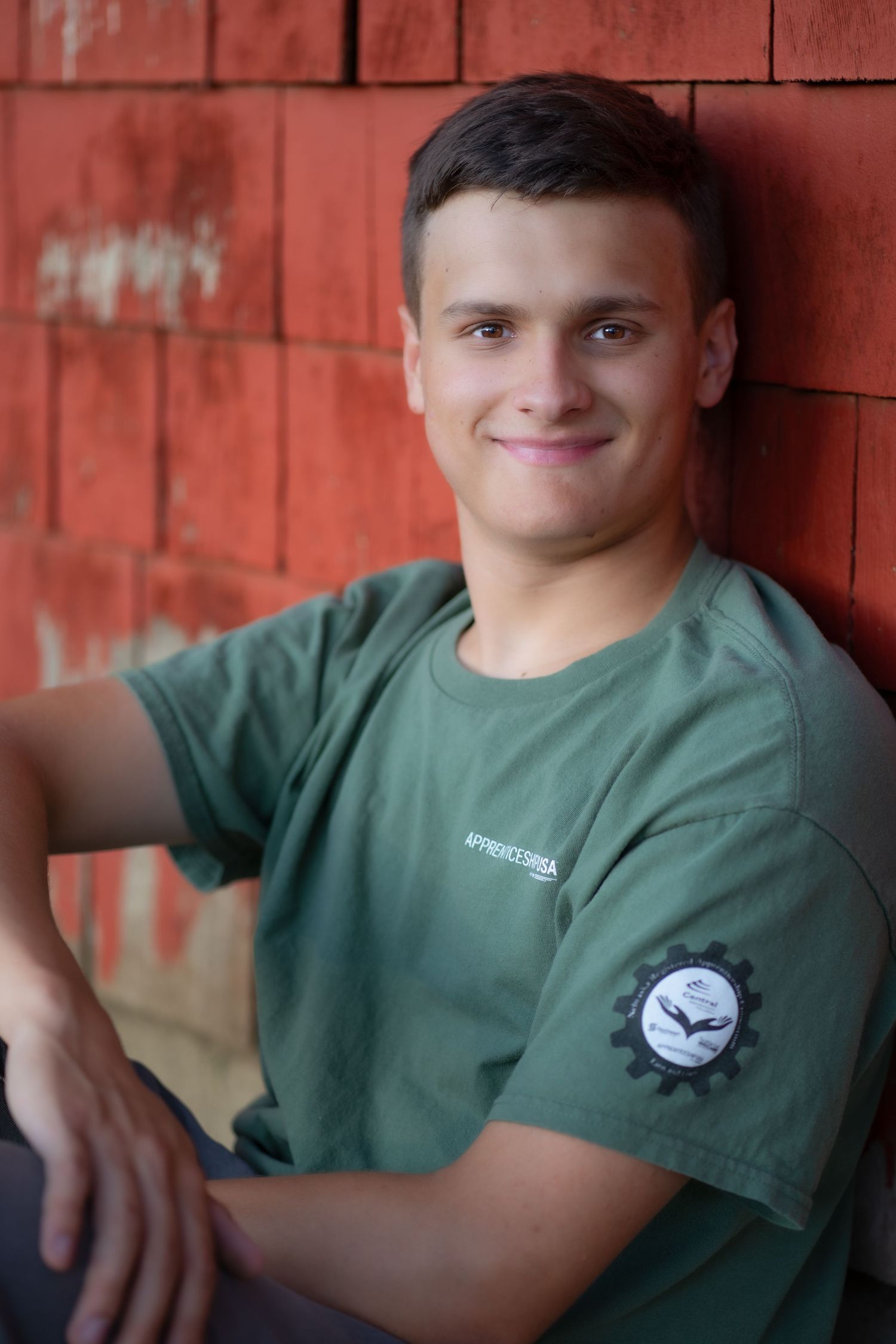 Smiling young person in green t-shirt with logo patch sits against rustic red brick wall in outdoor portrait.