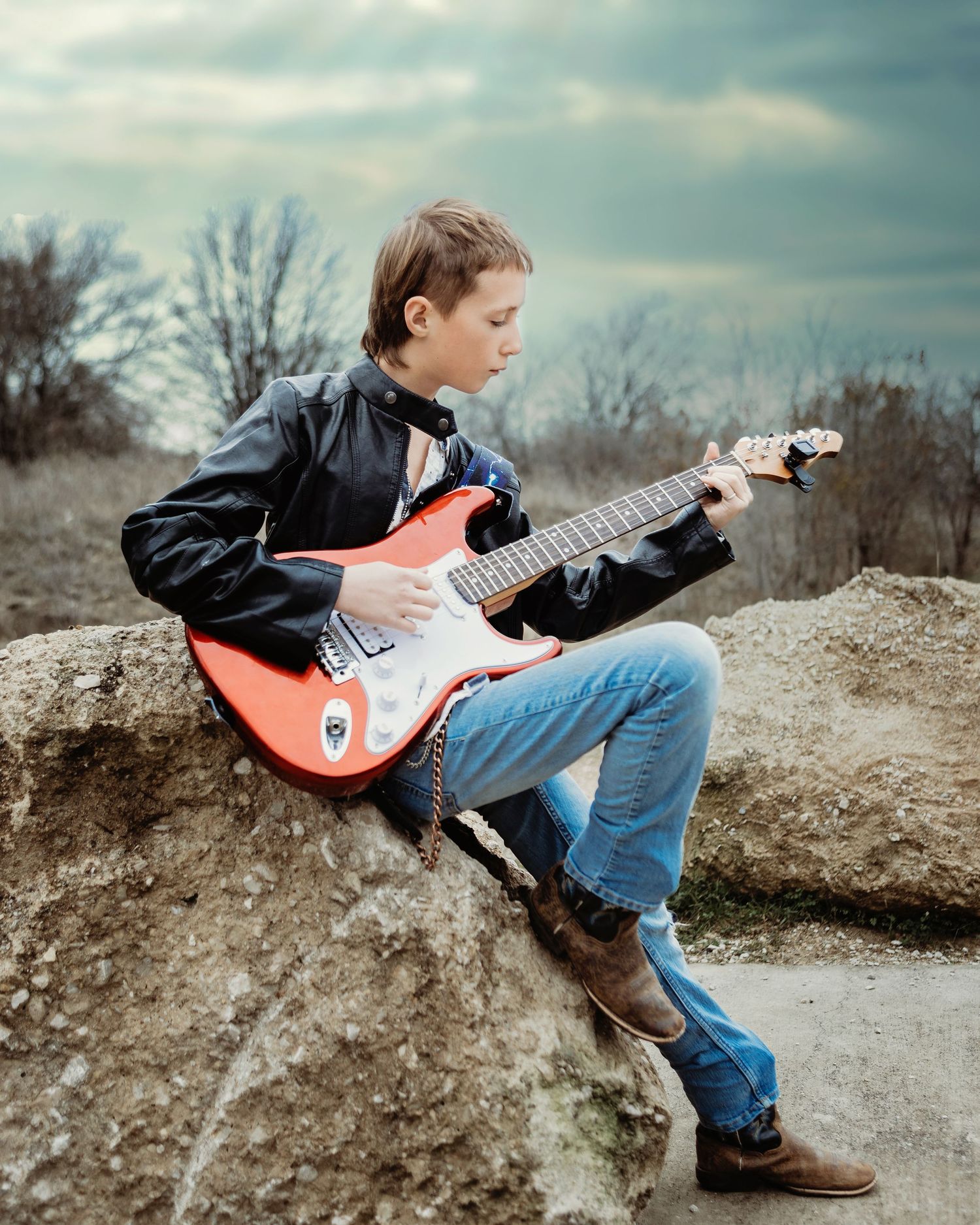 Boy playing a red electric guitar while sitting casually on a large rock against cloudy sky during photo session in Fort Worth, TX.