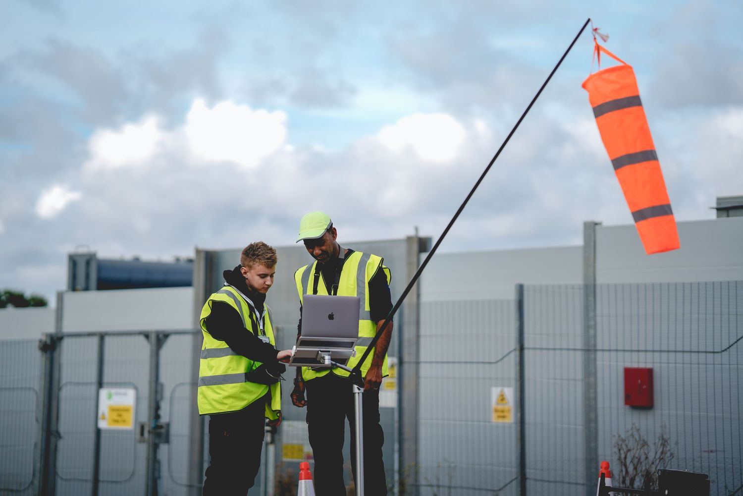 Two workers in safety vests measuring wind conditions with an orange windsock.