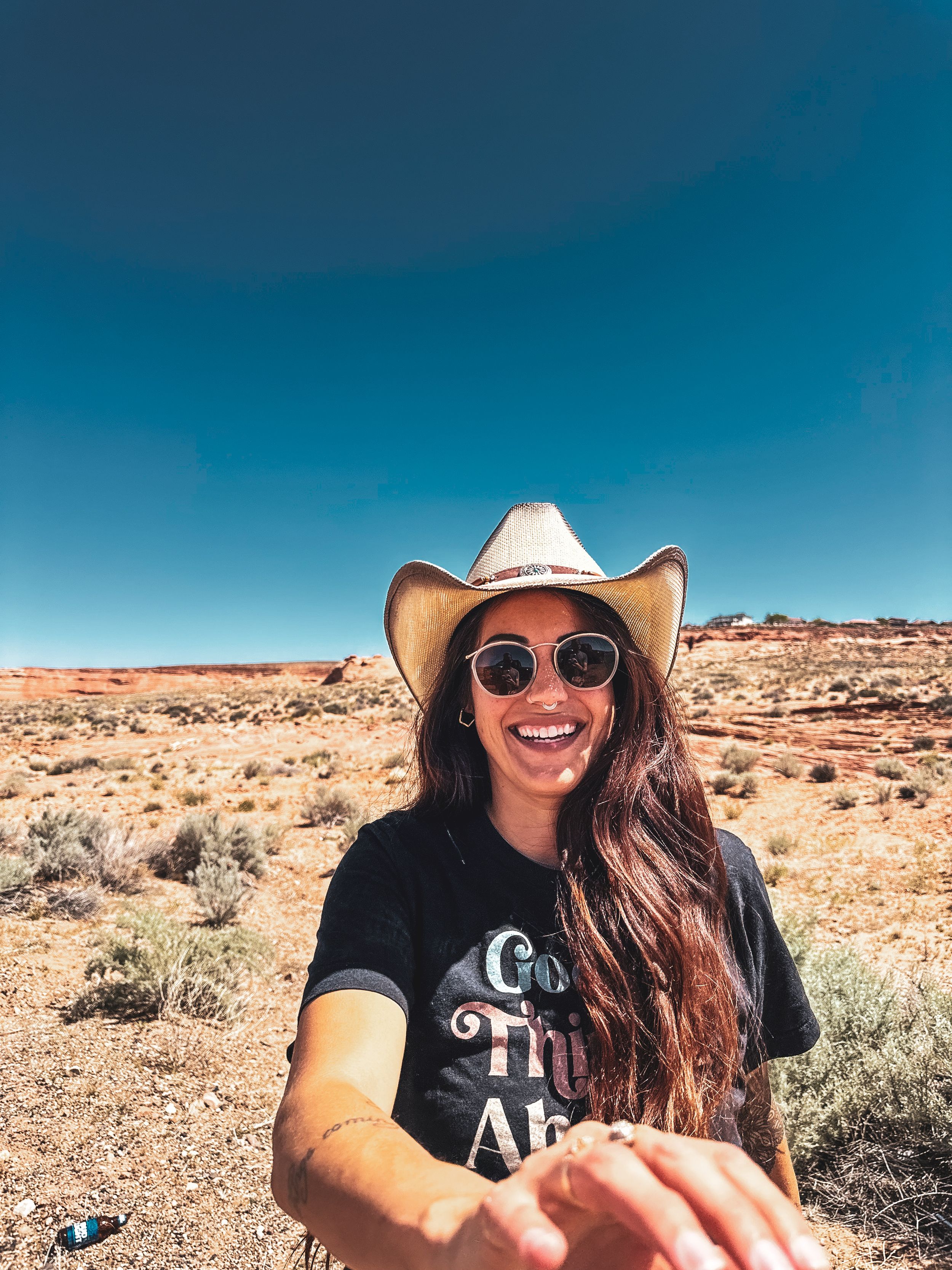 Smiling woman in cowboy hat and sunglasses in desert landscape selfie.