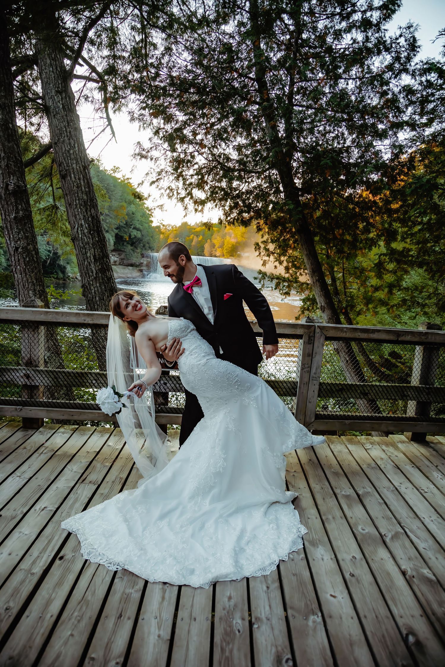 A romantic wedding couple dancing on a wooden deck surrounded by trees at sunset.