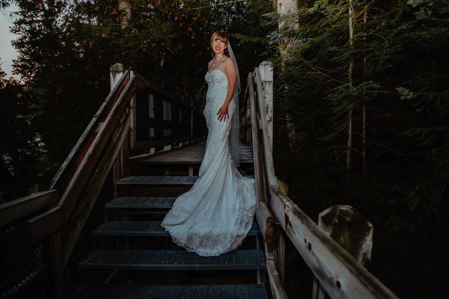 A bride in a flowing lace gown stands on wooden stairs in a forest setting.