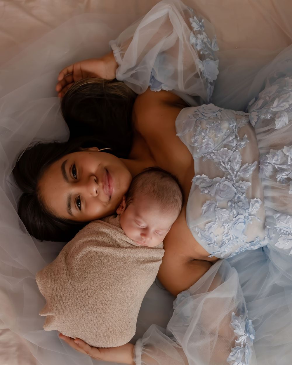 An artistic portrait of a mother in white lace dress lying with her sleeping newborn on tulle fabric.