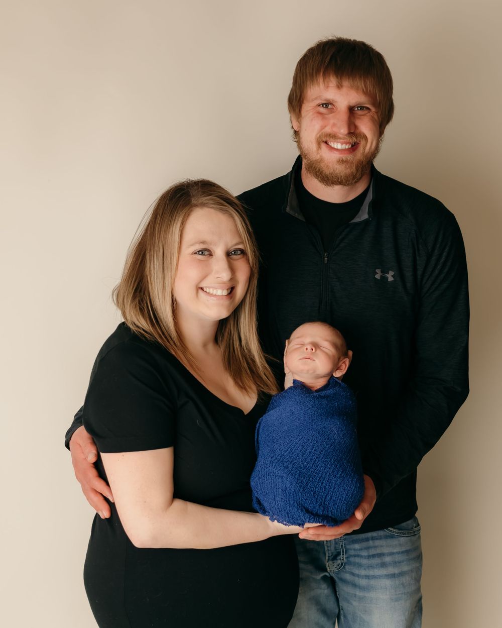 A studio portrait of new parents holding their sleeping baby wrapped in blue against a beige backdrop.