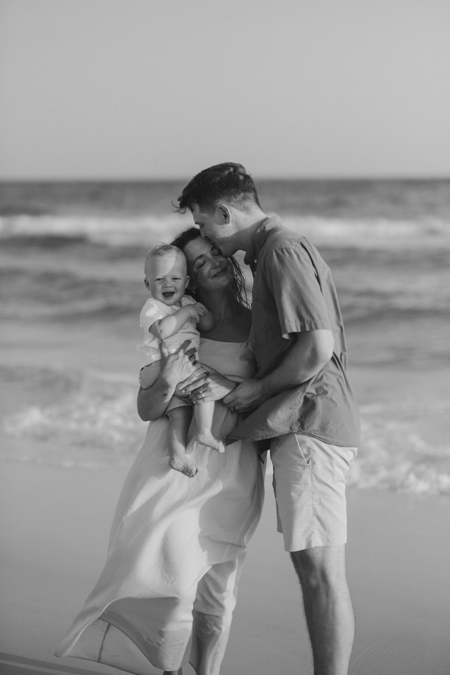 Black and white photos showing tender moments between parent and child playing on a beach with ocean waves.