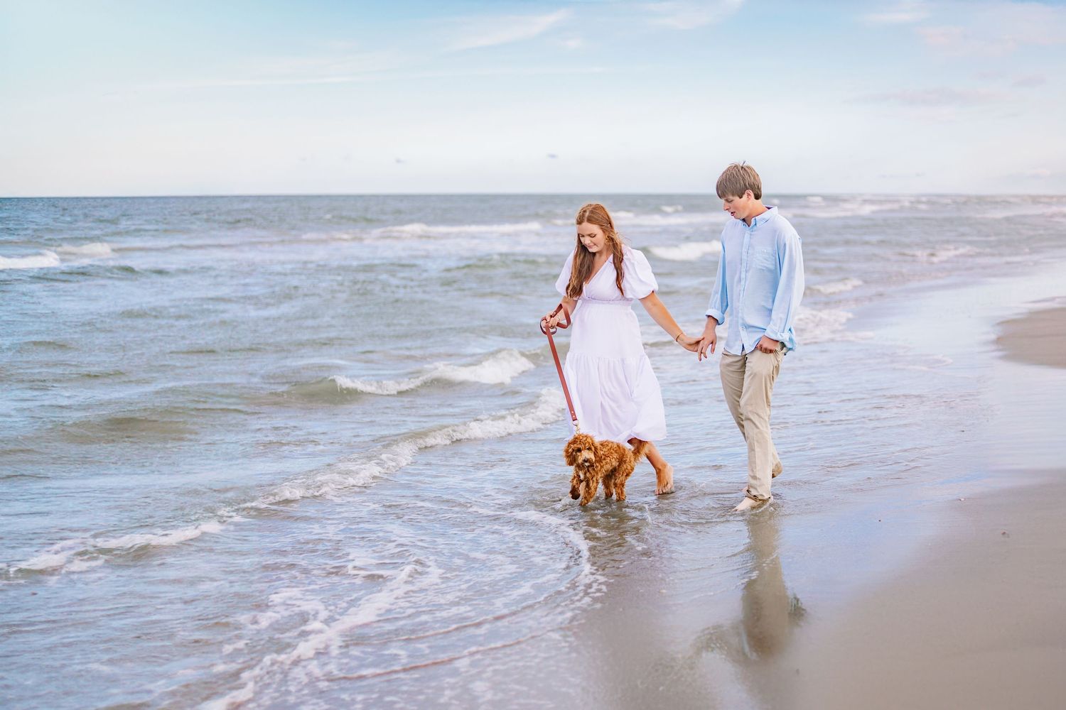 Couple plays with their fluffy brown dog along ocean shoreline at sunset.
