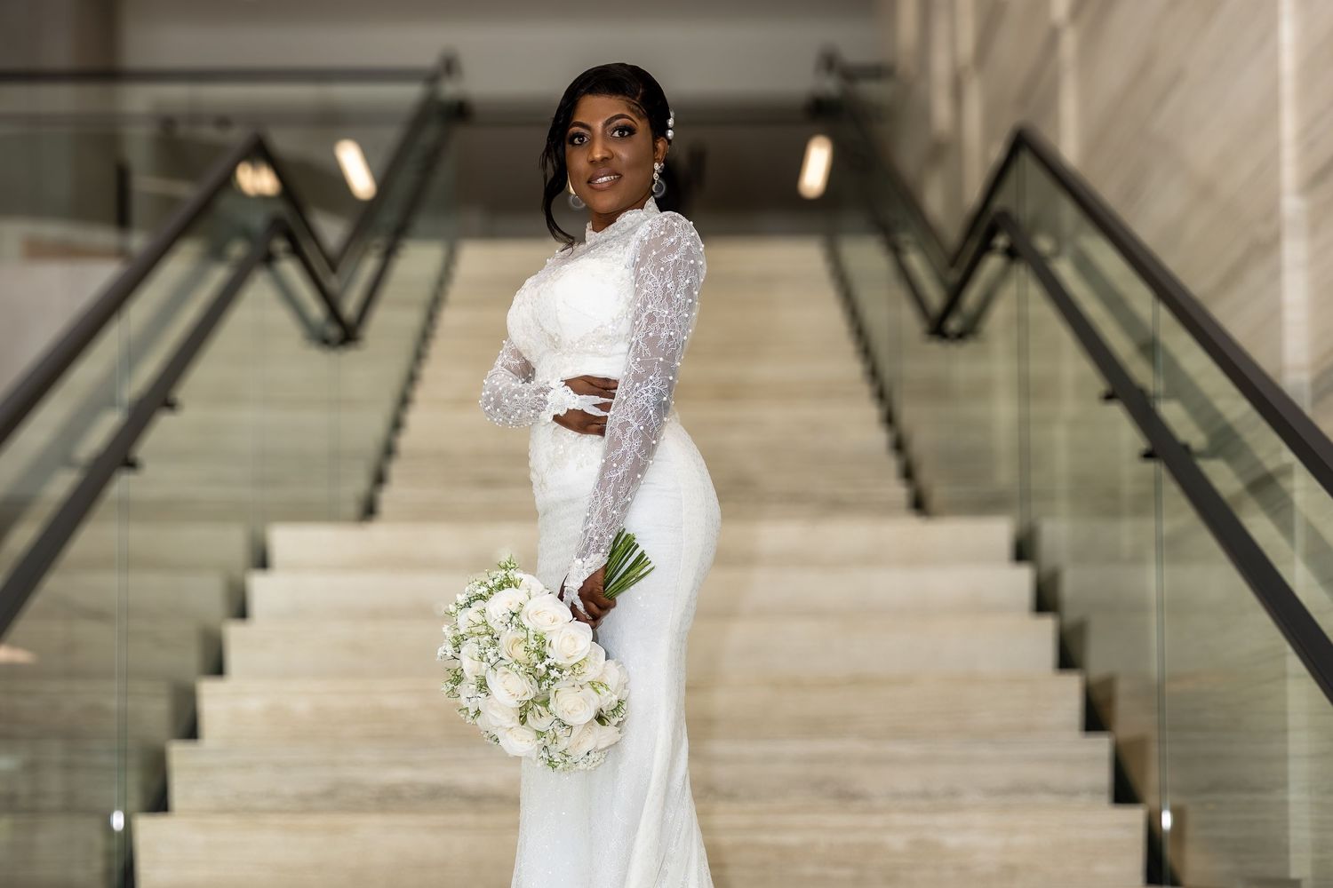 A bride in a white long-sleeved gown poses elegantly on marble stairs with a white bouquet.