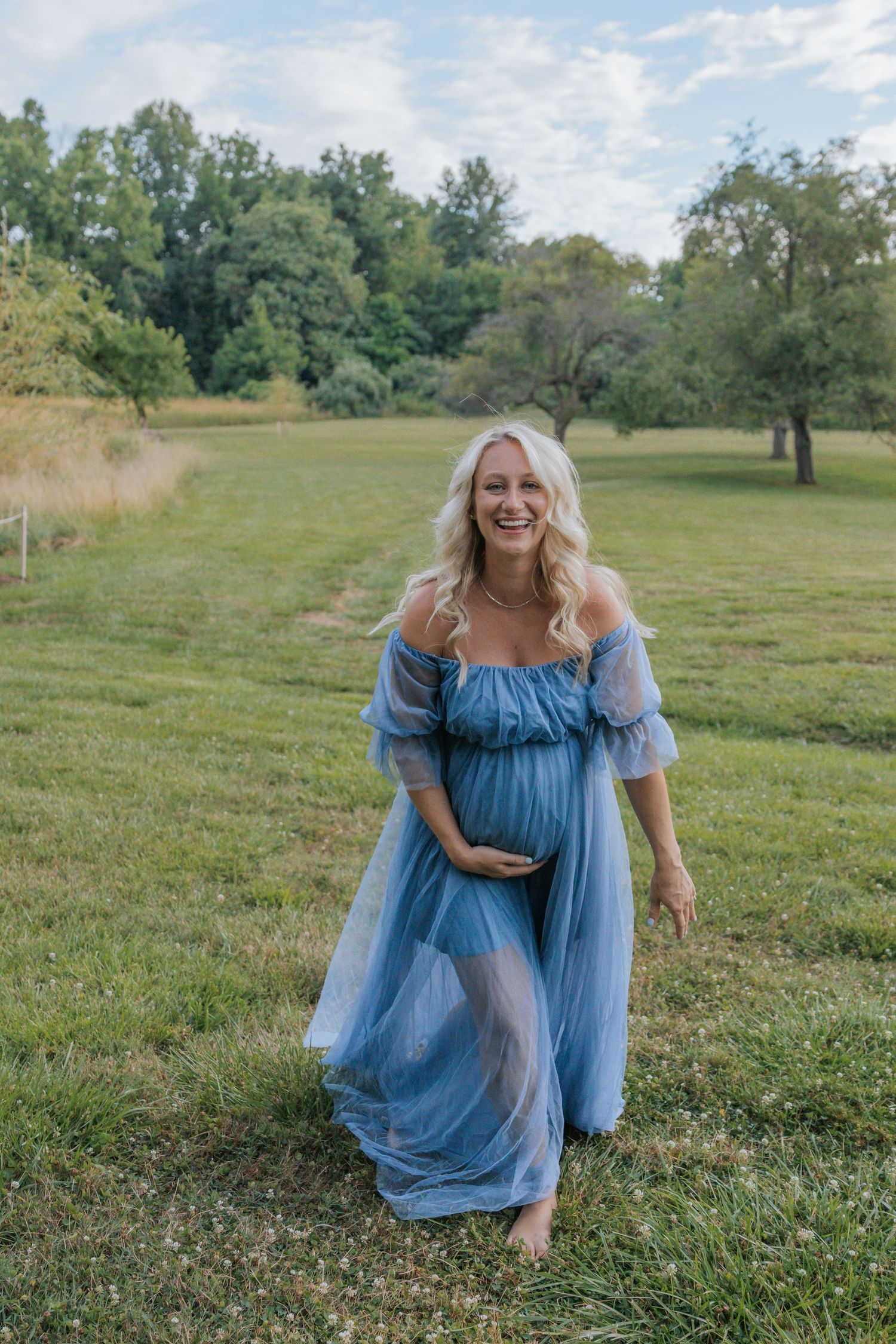 Expectant mother in flowing blue dress poses in sunny meadow.