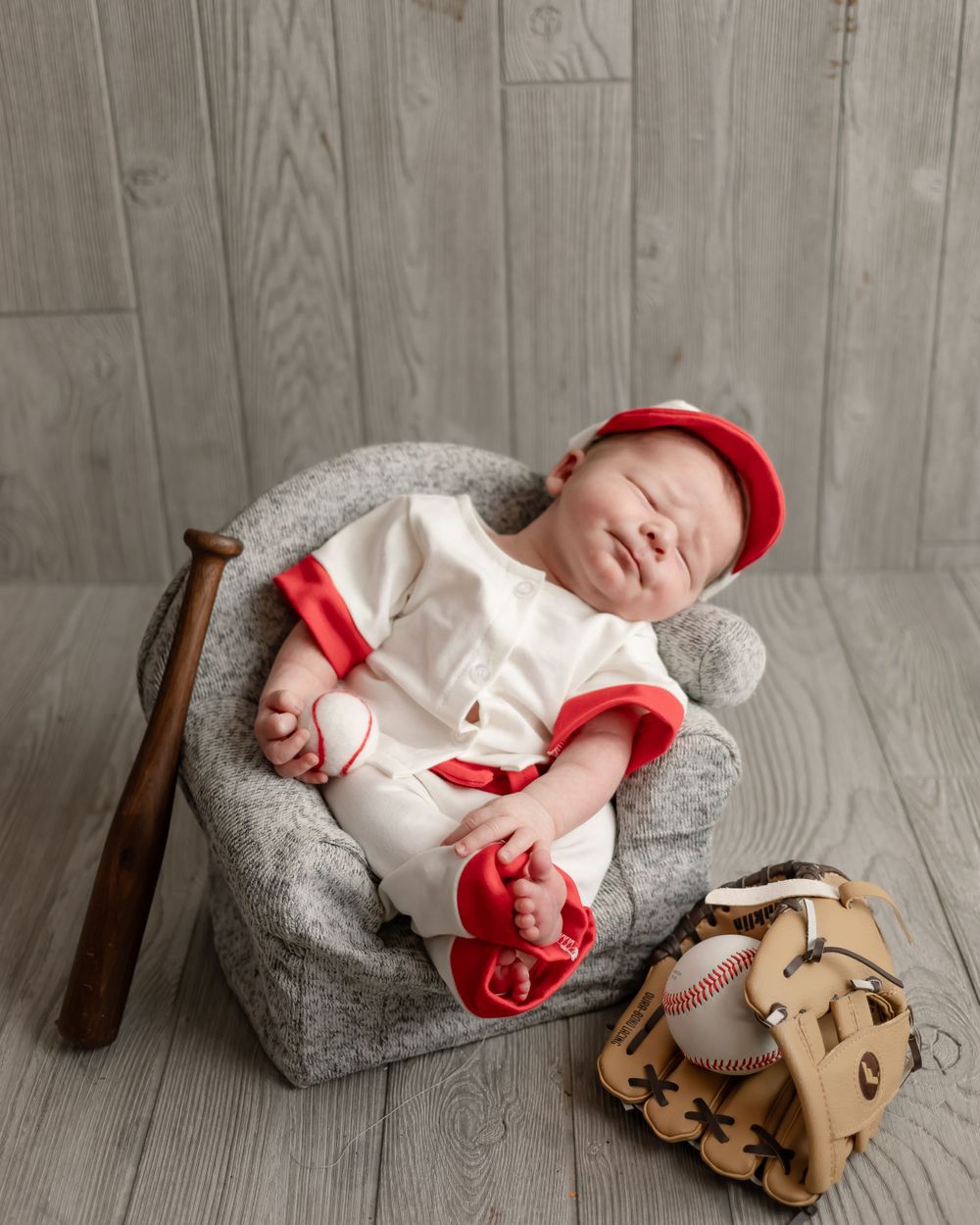 Sleeping baby in baseball outfit with red trim posed with mitt and ball on gray backdrop.