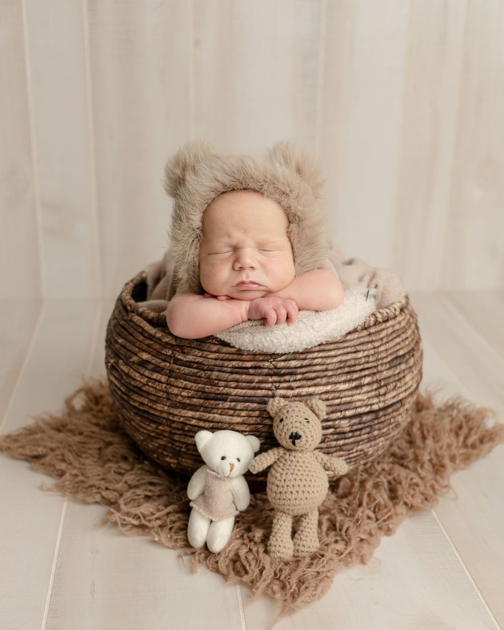 Sleeping newborn in furry hat rests in woven basket with stuffed bear toys on brown textured blanket.