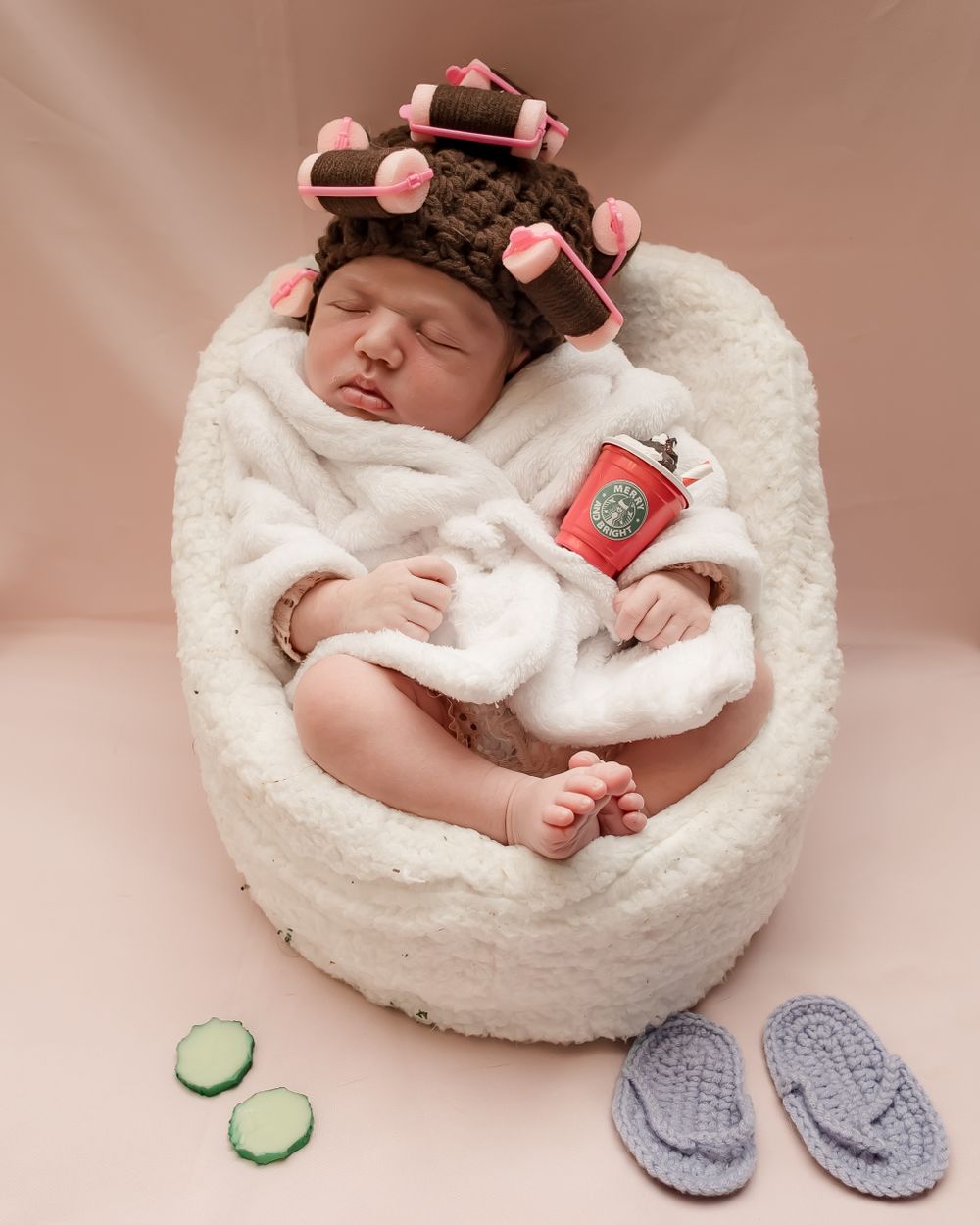 Newborn relaxes in plush chair wearing pink curlers and holding tiny coffee cup with cucumber slices nearby.