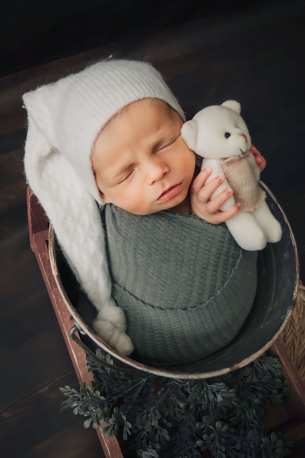 Sleeping newborn wrapped in grey knit blanket holds white stuffed animal in wooden basket.