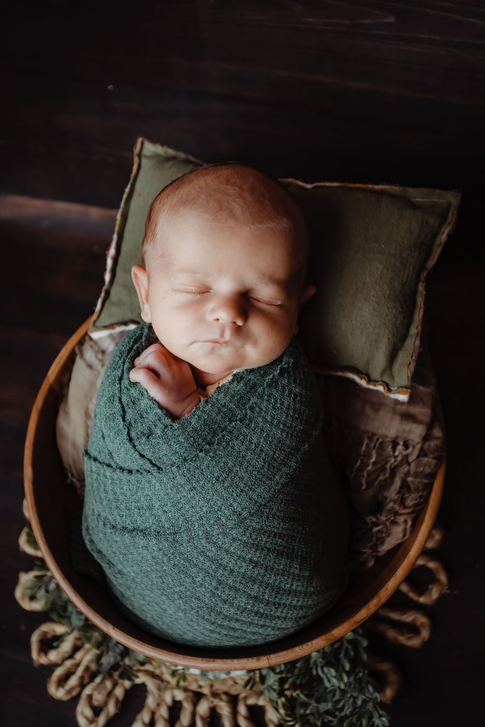 Peaceful newborn baby swaddled in dark green wrap sleeping in wooden bowl.