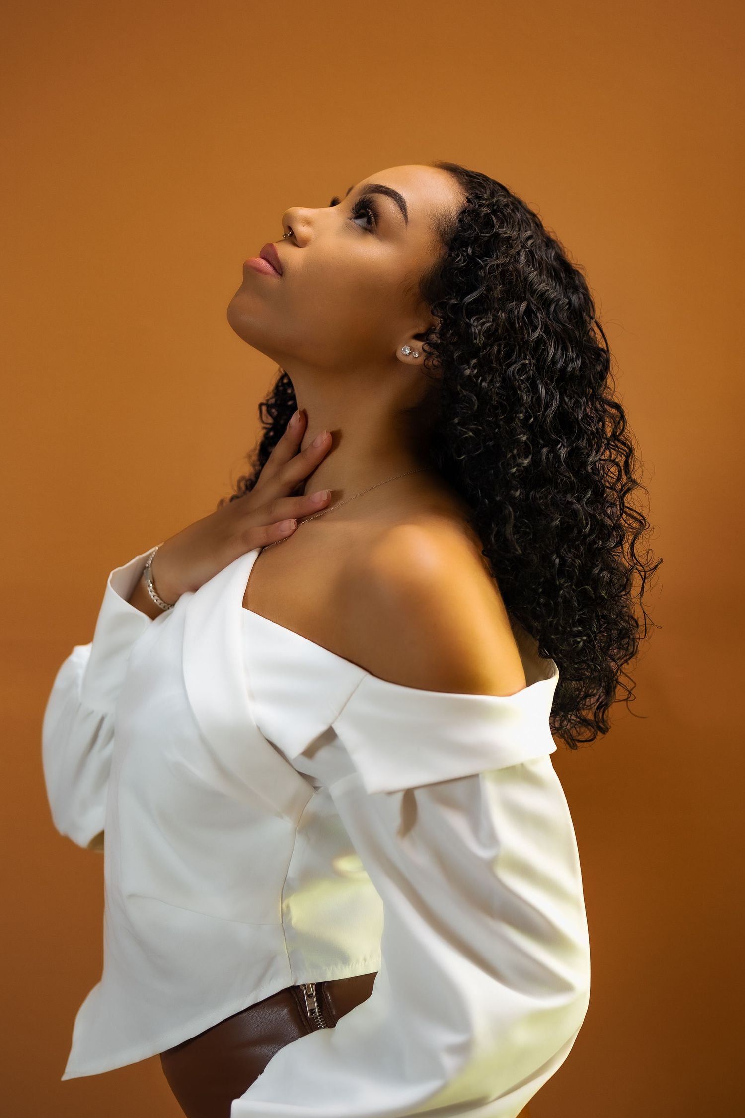 Artistic profile portrait against warm orange background wearing white off-shoulder top.