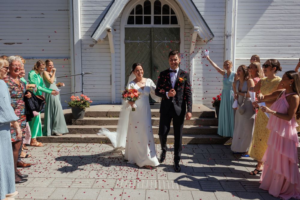 A newlywed couple exits a white church while guests shower them with flower petals.