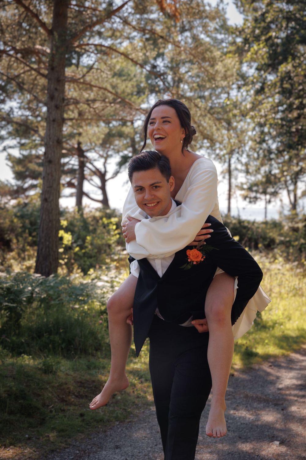 A playful piggyback ride moment between a couple in elegant wedding attire surrounded by pine trees.