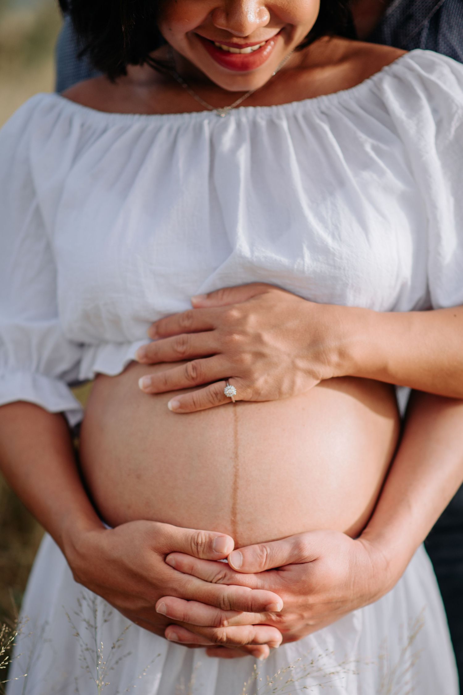 Pregnant woman in white dress tenderly touching her baby bump while wearing an engagement ring.