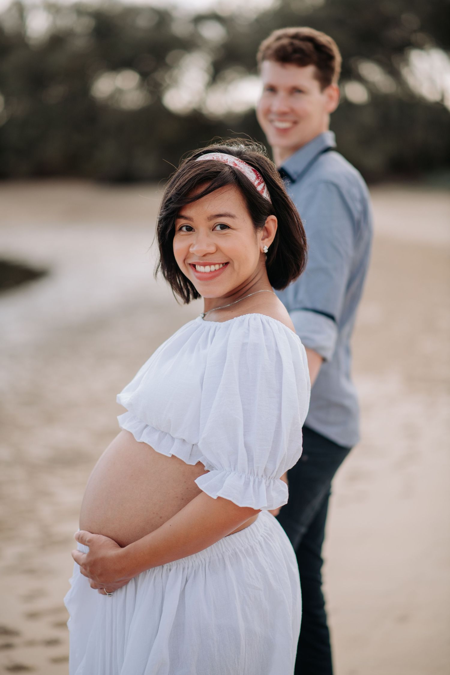 Couple poses for maternity photo on beach at sunset with woman in flowing white dress.