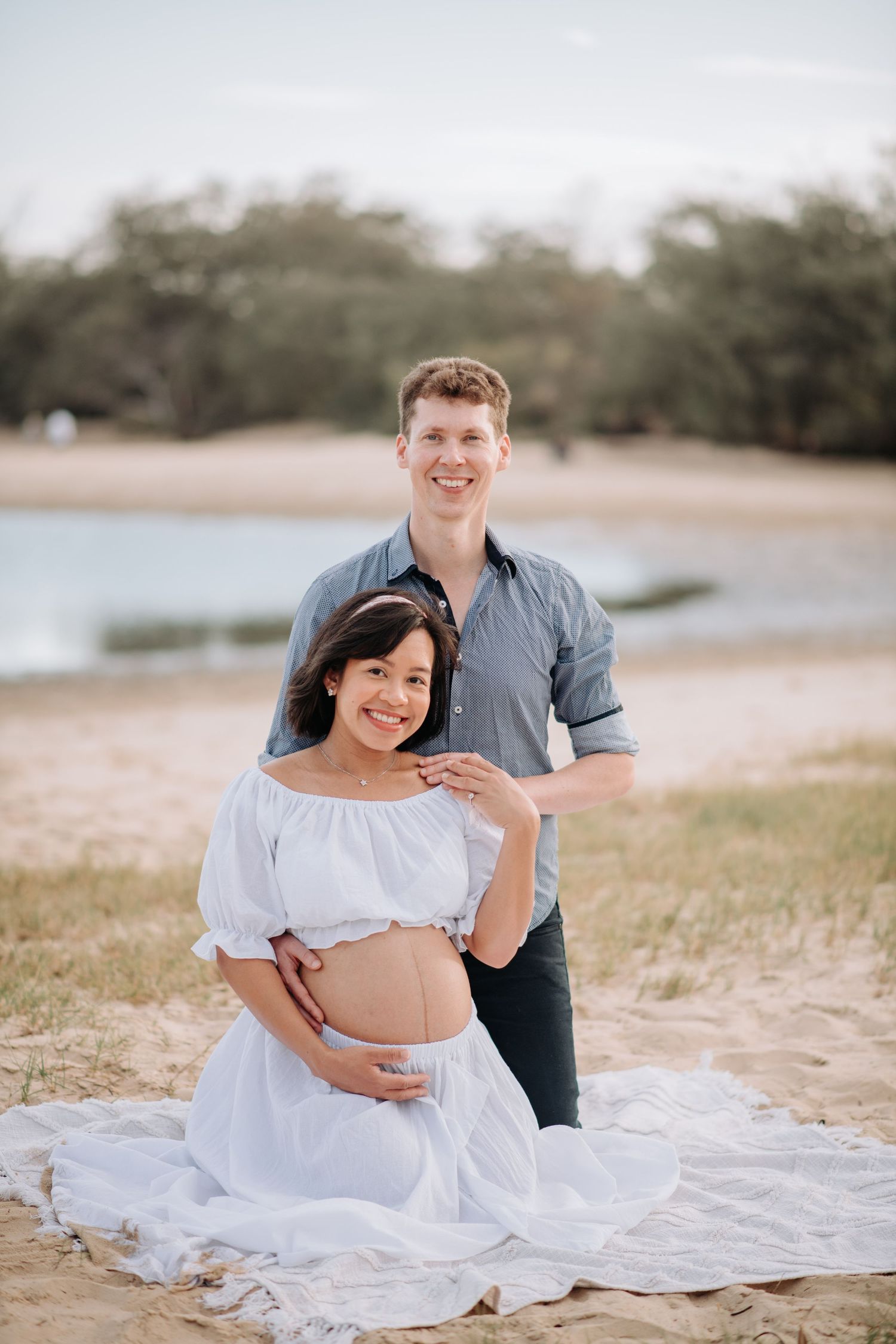 Expecting couple poses on beach blanket during sunset maternity photoshoot.