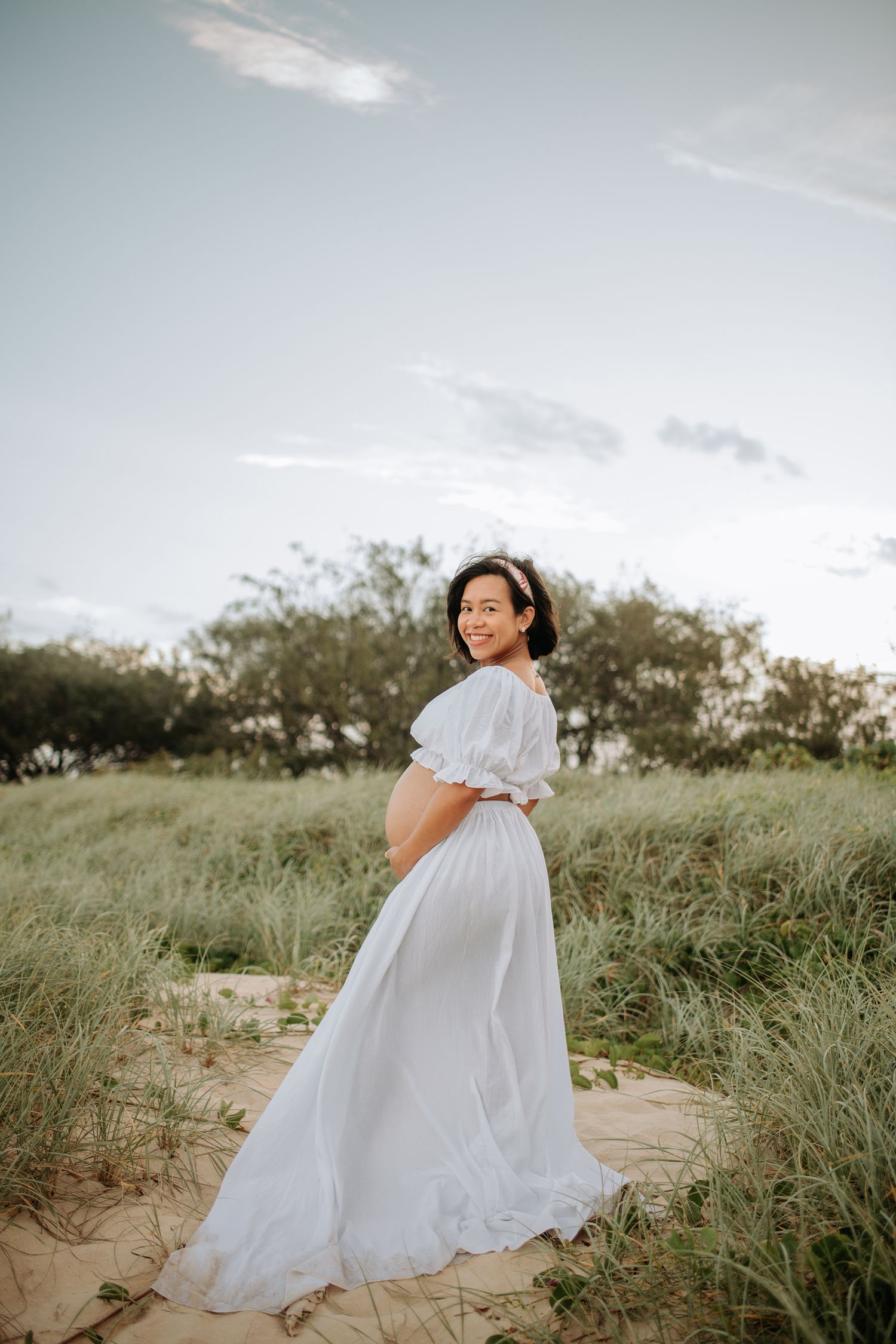 Ethereal maternity portrait of woman in flowing white dress on sandy dunes at sunset.