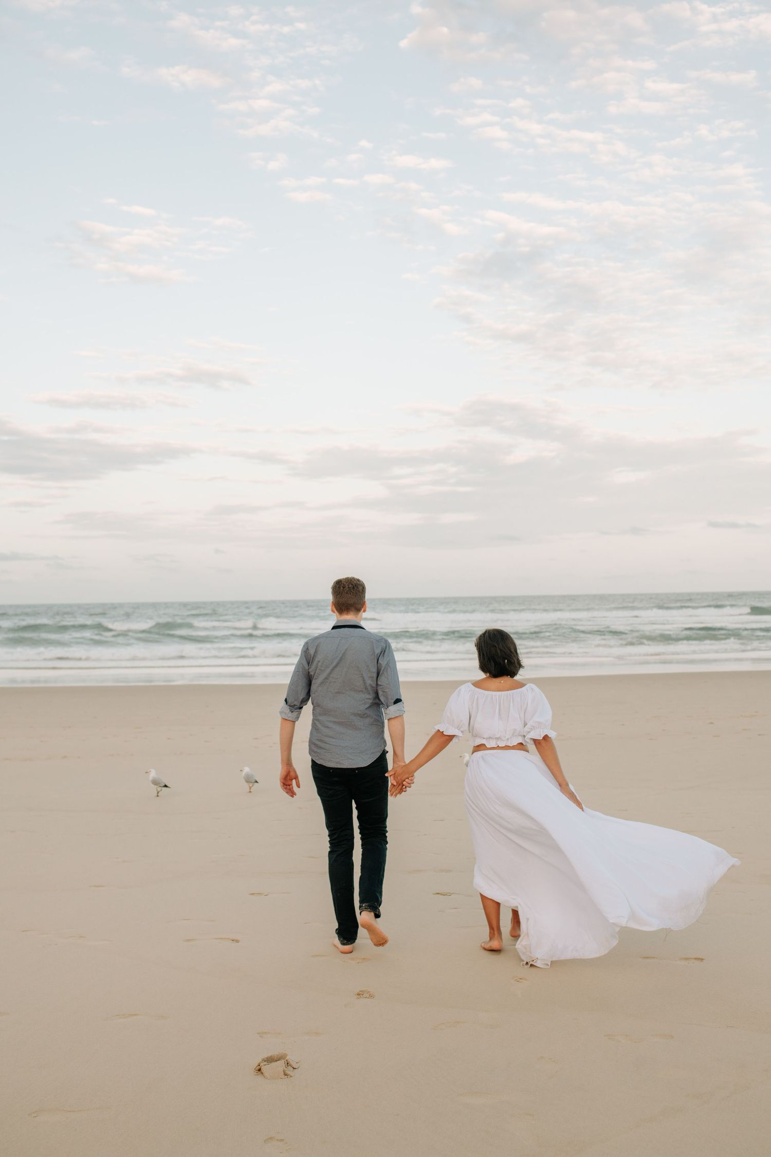 Couple walking hand in hand on empty beach at sunset with flowing white dress.