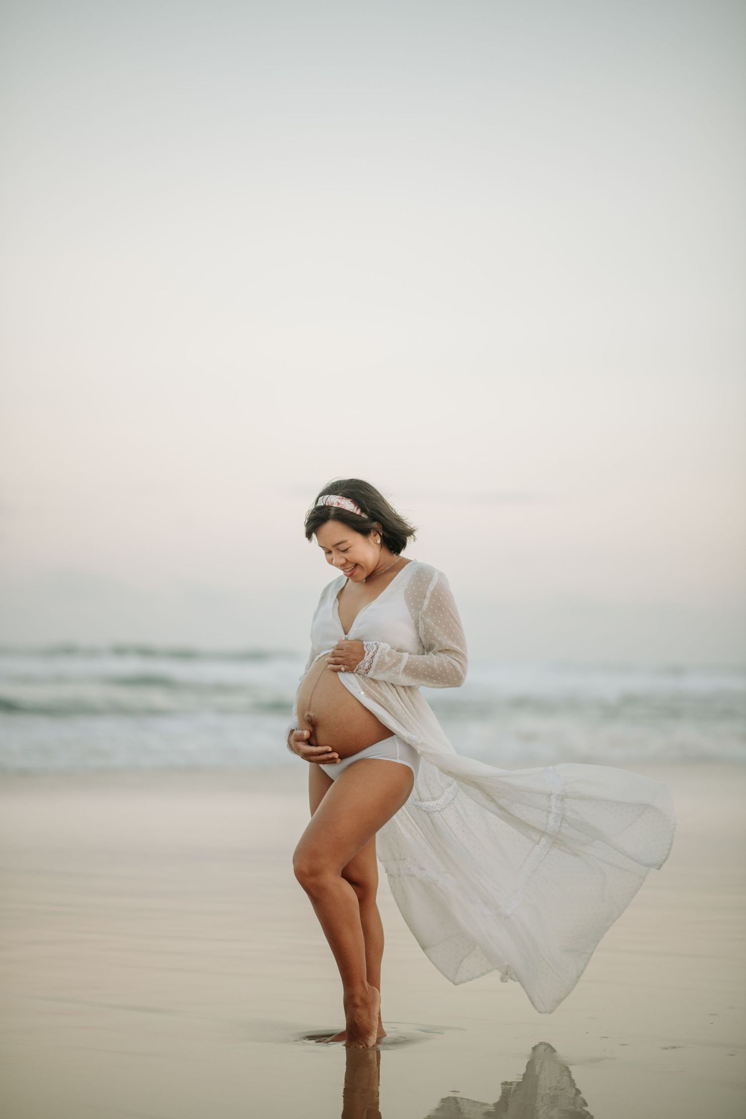 Ethereal maternity photo of woman in flowing white dress standing on beach at sunset.
