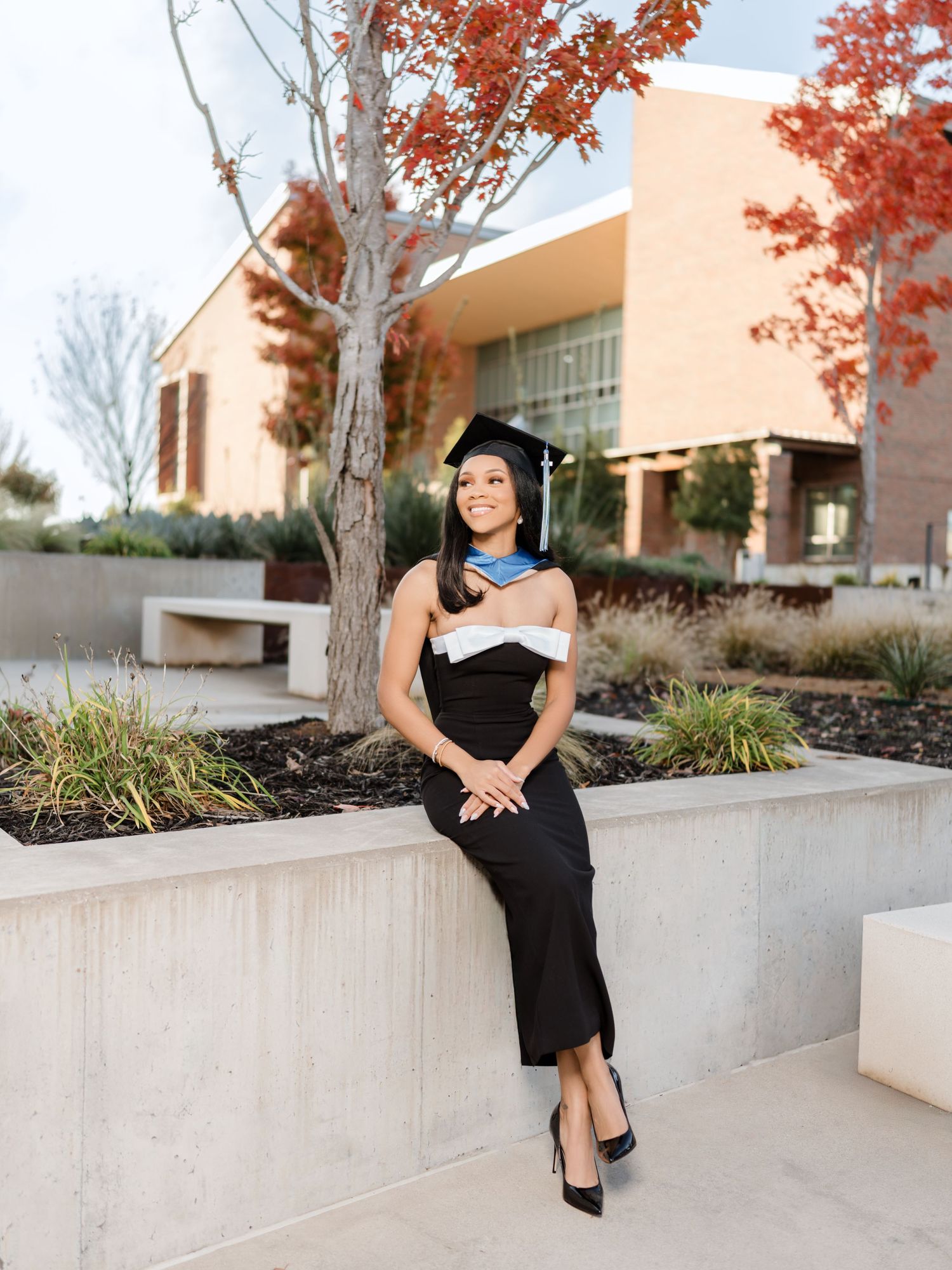 Graduate in cap and gown poses on concrete ledge with autumn trees and building in background.
