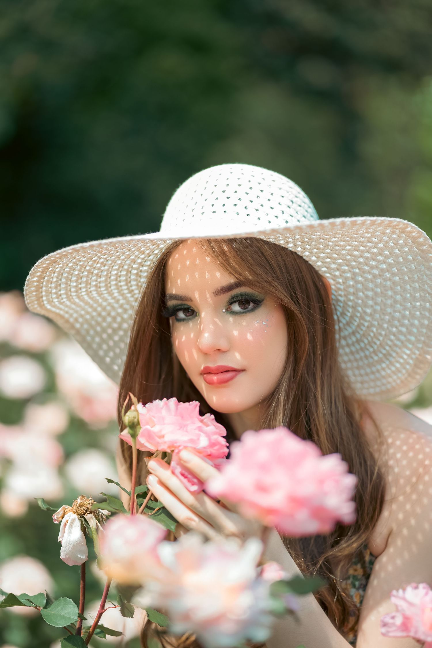 Woman in white sunhat poses among blooming pink roses in garden setting.