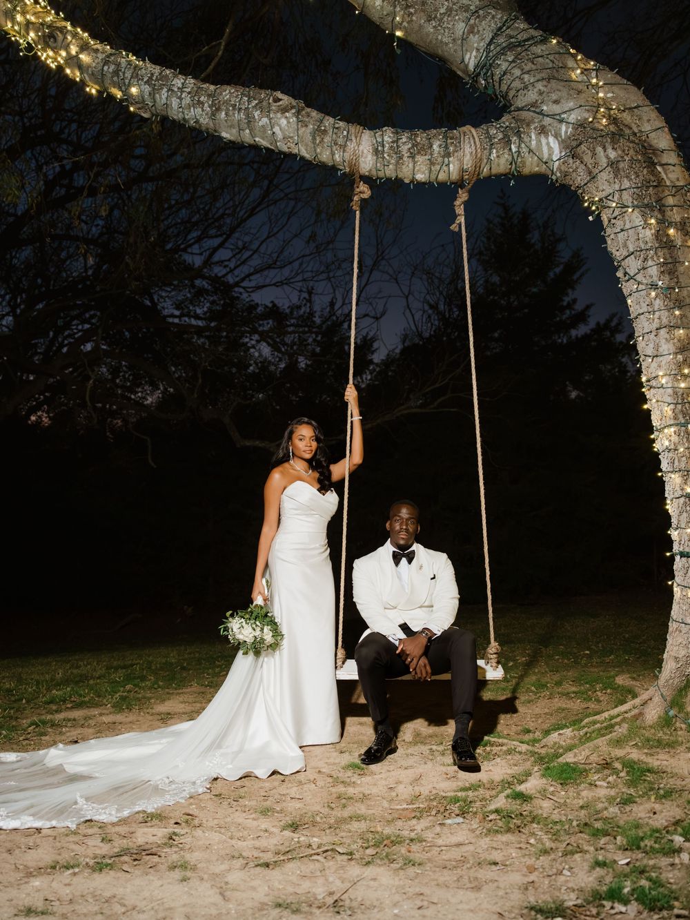 Romantic wedding couple swings together on tree swings at dusk in a dreamy outdoor garden setting.