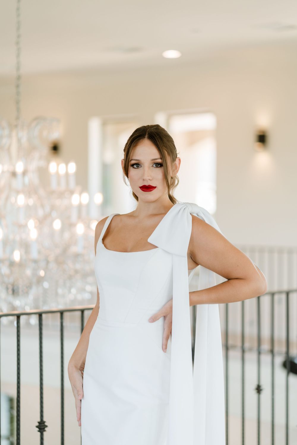 Elegant bride in white dress with bow detail poses against chandelier backdrop.