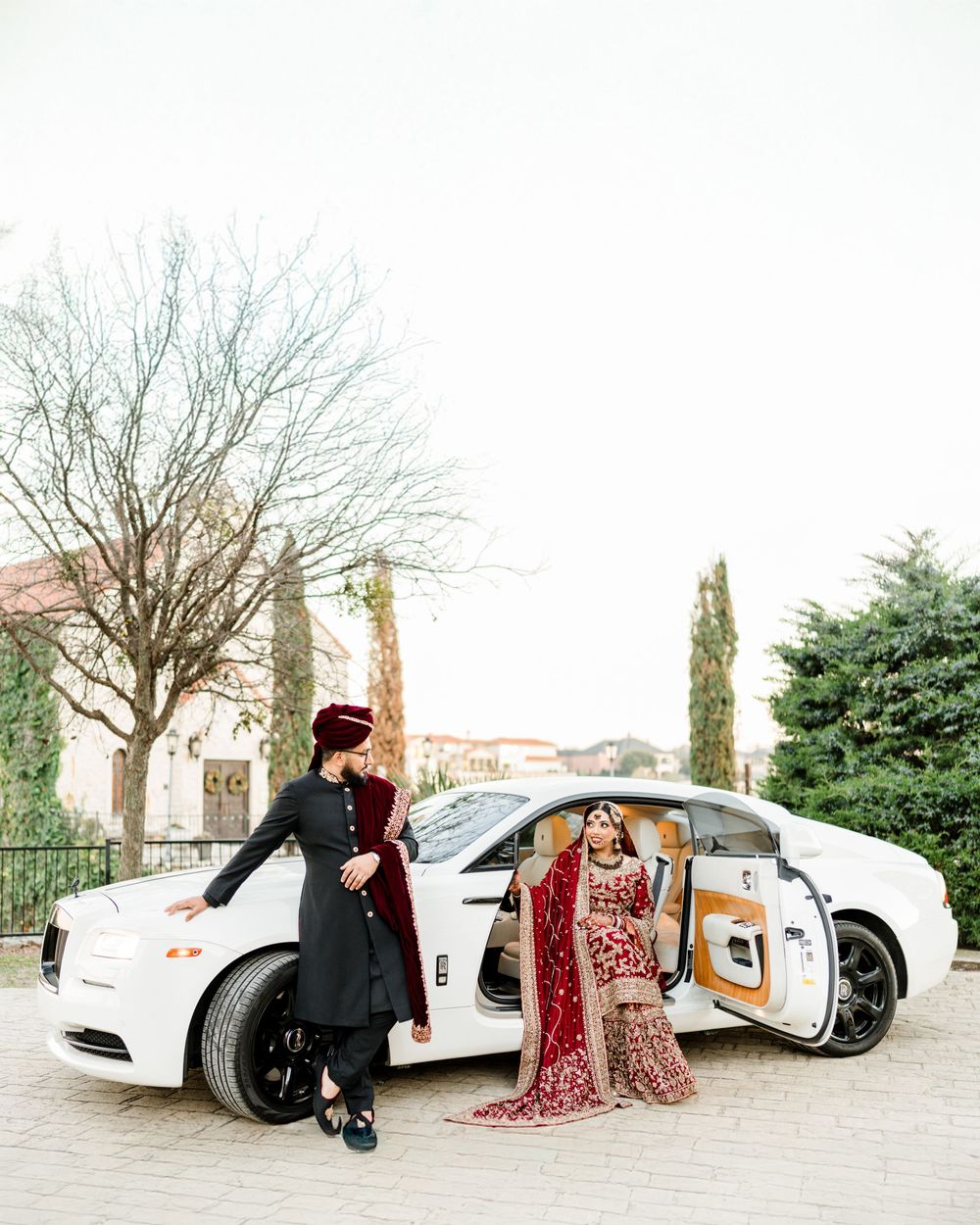 Luxury white car parked near historic building with people dressed in traditional wedding attire standing beside it.