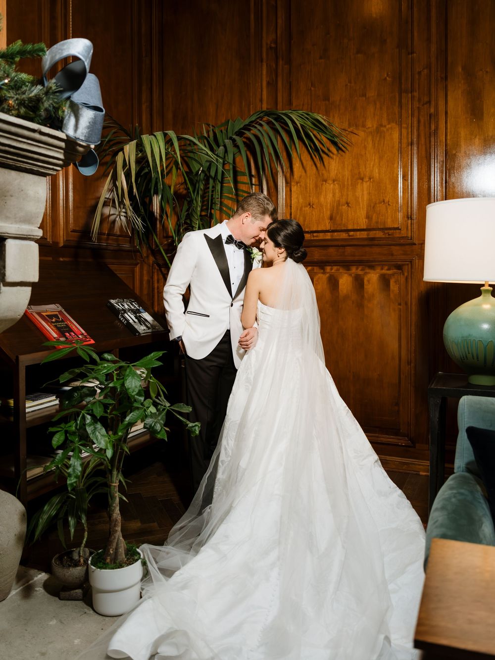 Elegant wedding couple shares intimate moment in wood-paneled room with tropical plants and vintage decor.