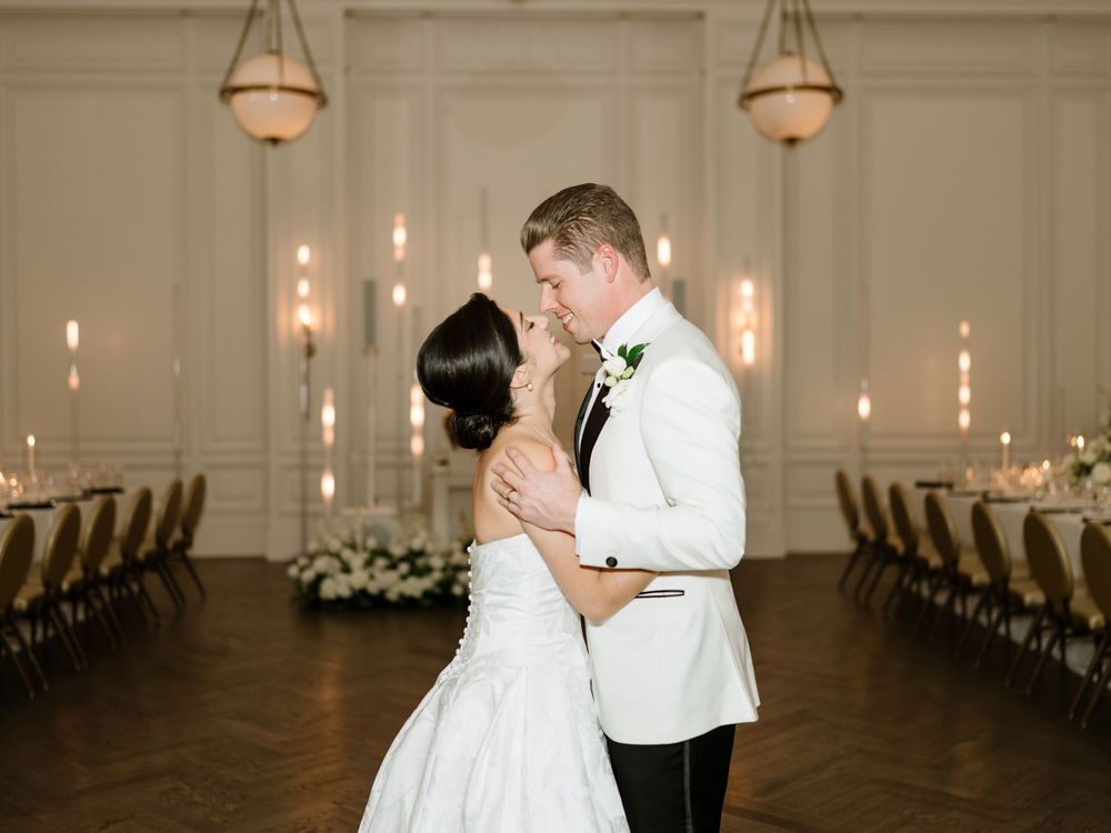 Newlyweds share romantic moments in an elegant ballroom with soft lighting and white floral arrangements.
