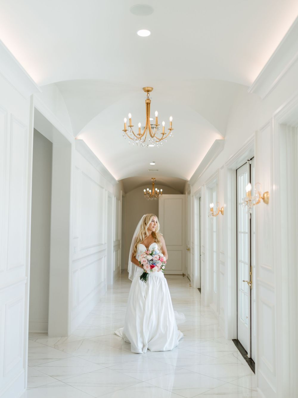 Elegant white hallway with crystal chandeliers and classic molding features a bridal gown displayed at the far end.