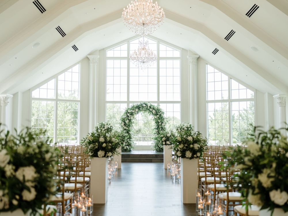 Elegant white chapel interior with crystal chandeliers, floral arch and rows of wooden chairs for a wedding ceremony.