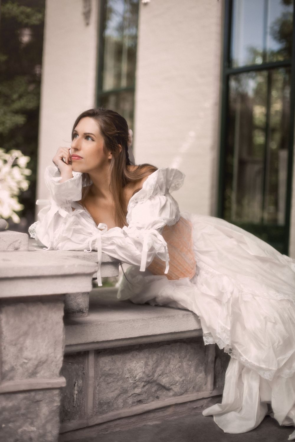 Fantasy session in a flowing white gown with puffed sleeves, leaning thoughtfully on a stone ledge in a soft, dreamy outdoor setting