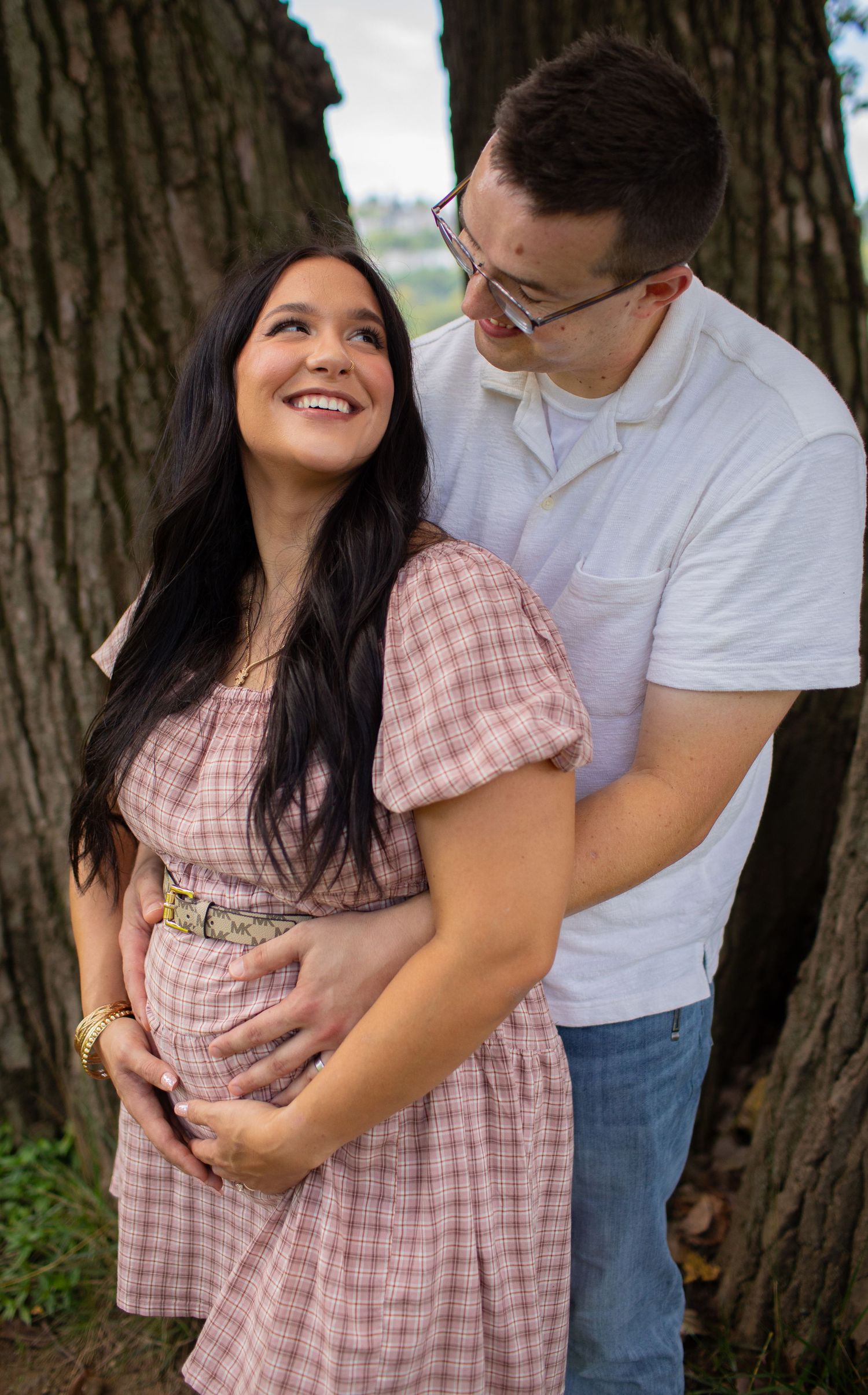A smiling pregnant woman in a pink dress embraces her partner near large trees outdoors.