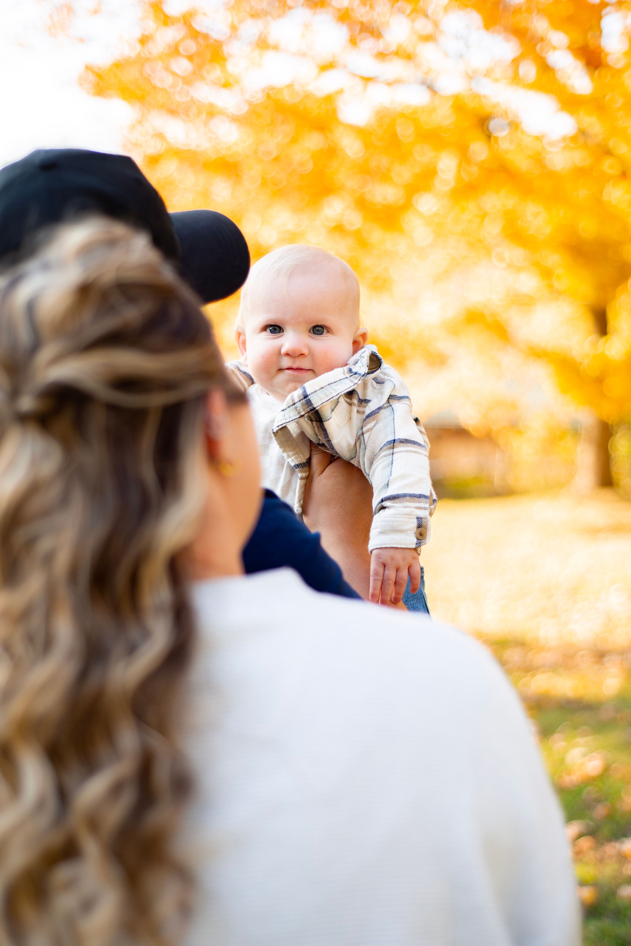 A baby in a plaid shirt is held up by parents during an autumn family photo session with golden foliage.