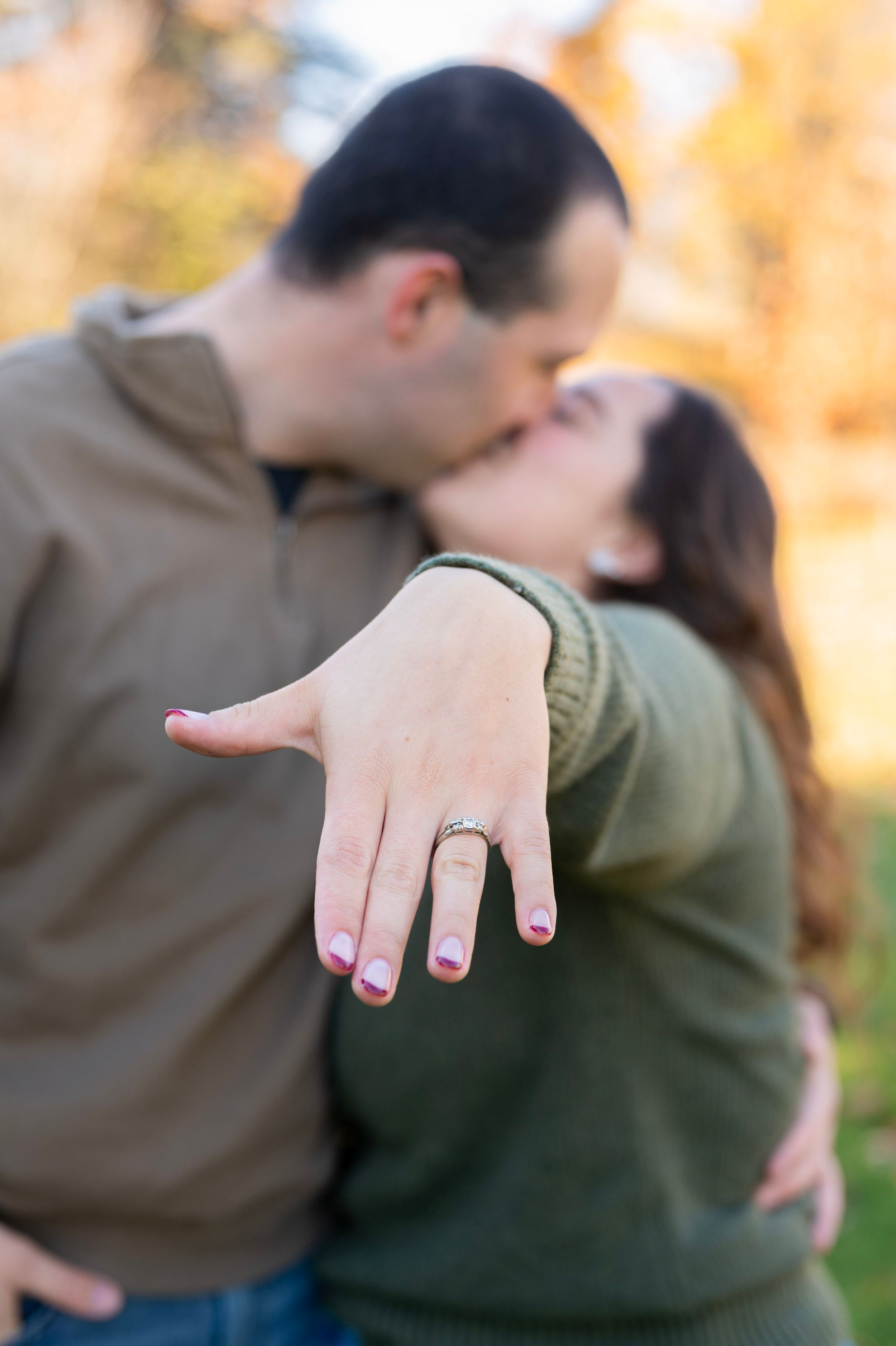 Couple sharing a kiss while showing off an engagement ring with autumn foliage in the background.