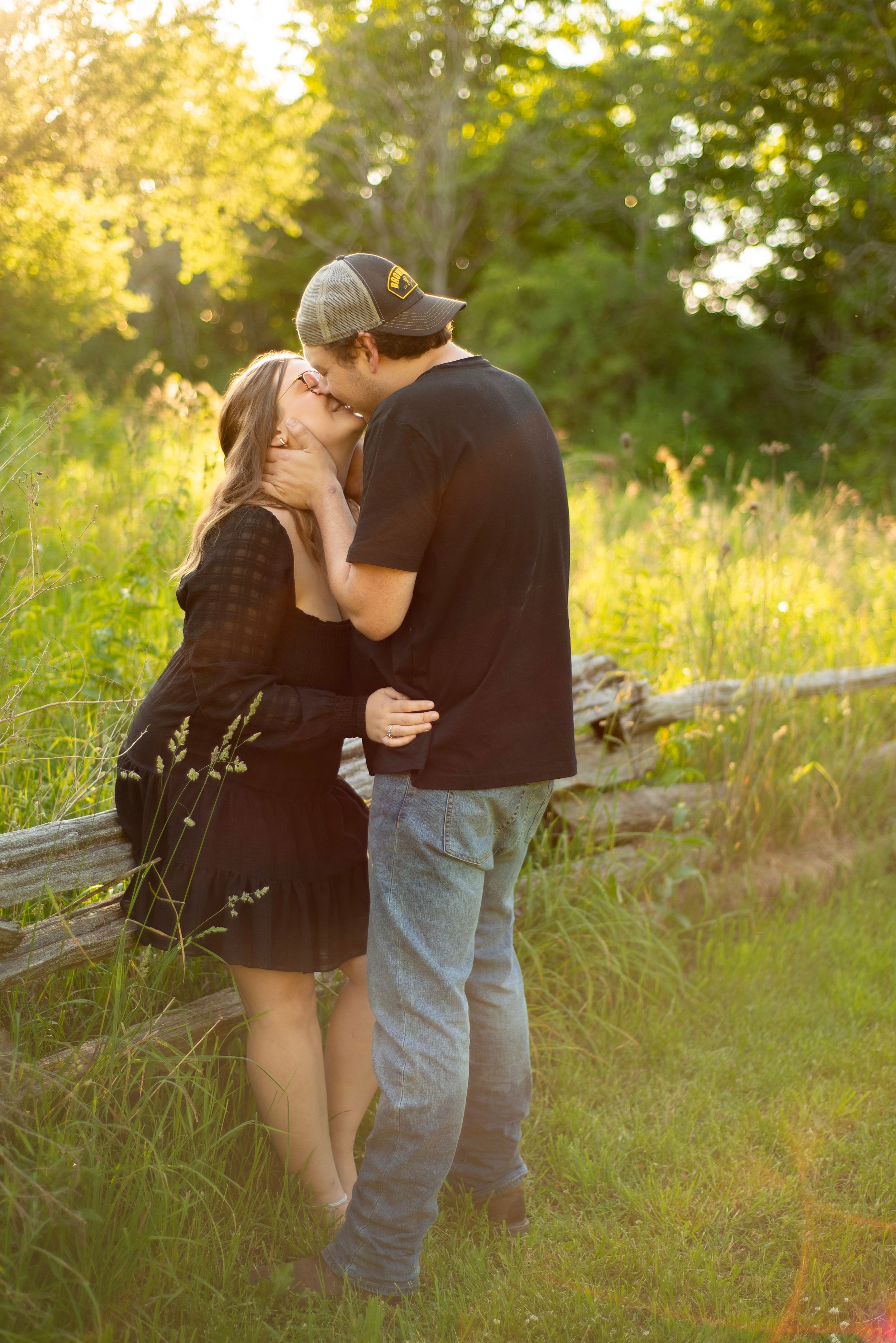 Romantic couple embracing in a sunlit meadow beside a rustic wooden fence at golden hour.