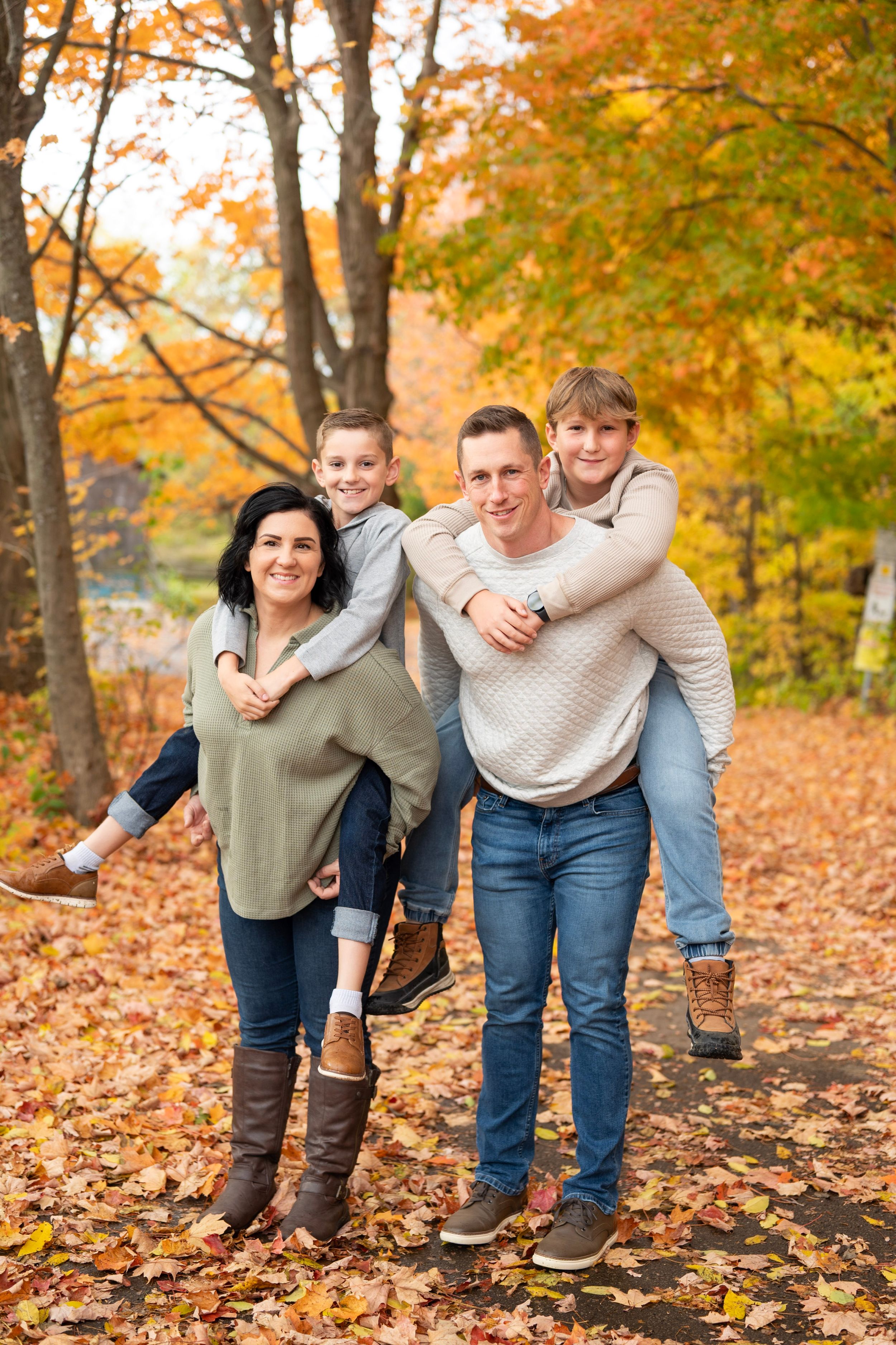 Family of four enjoying autumn piggyback rides on a leaf-covered path surrounded by colorful fall foliage.