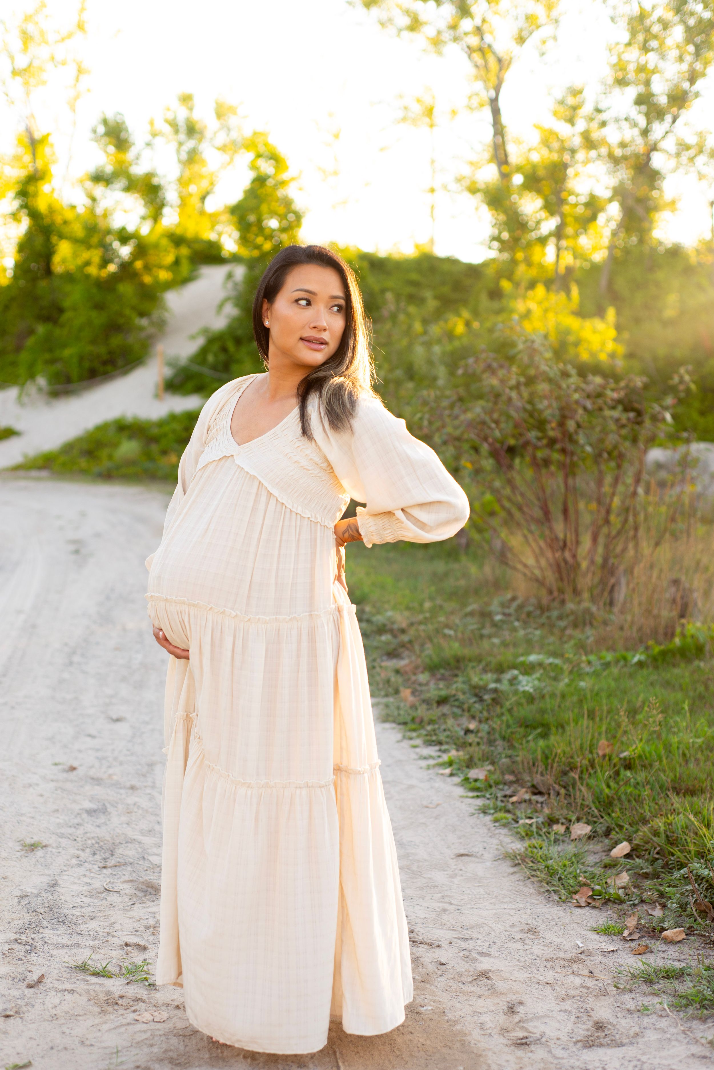 Pregnant woman in cream maxi dress standing on sandy path at golden hour with lush green trees in background.