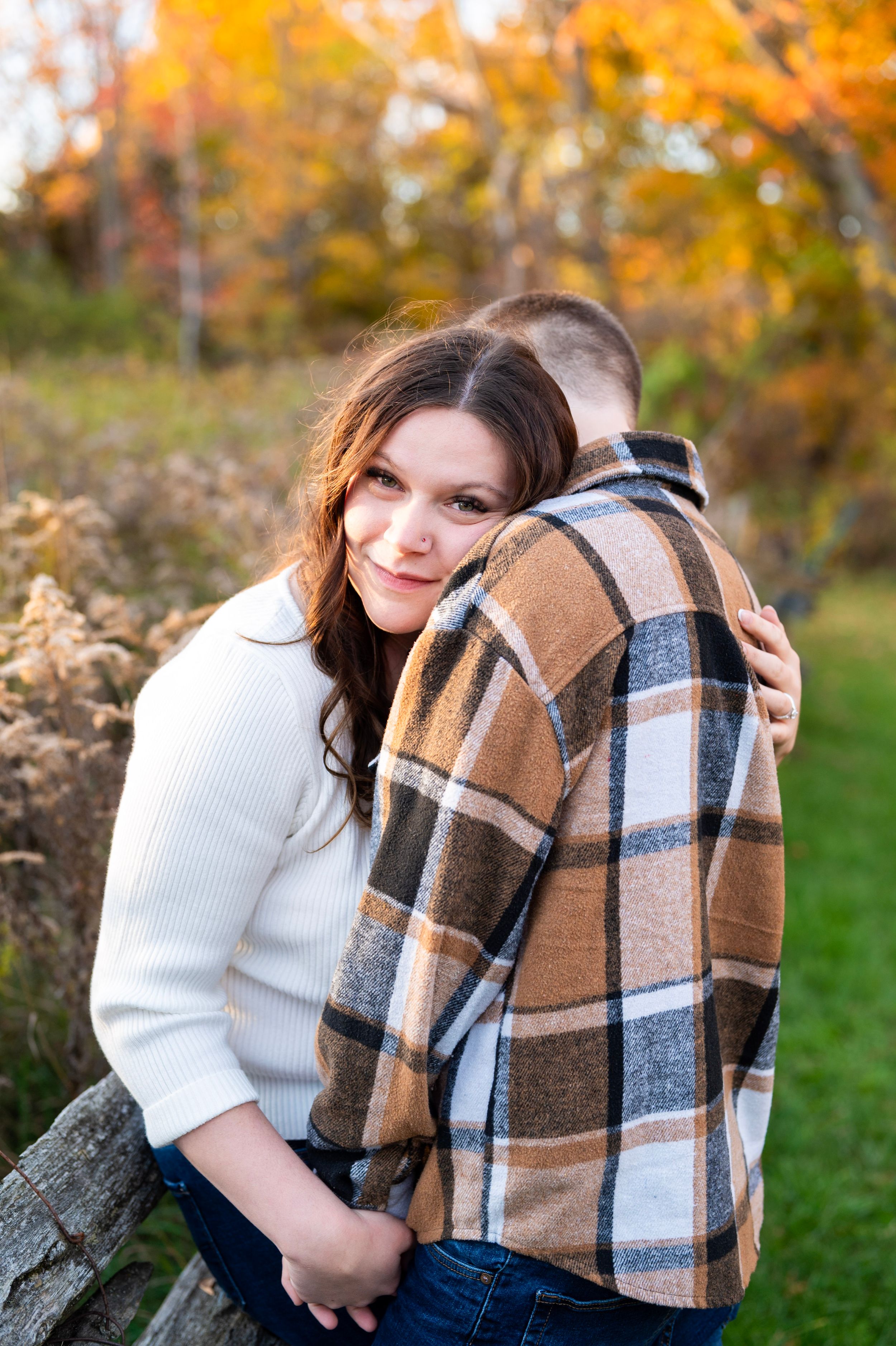 Couple embracing outdoors in autumn, woman in white sweater hugging man in plaid shirt, colorful fall foliage background.