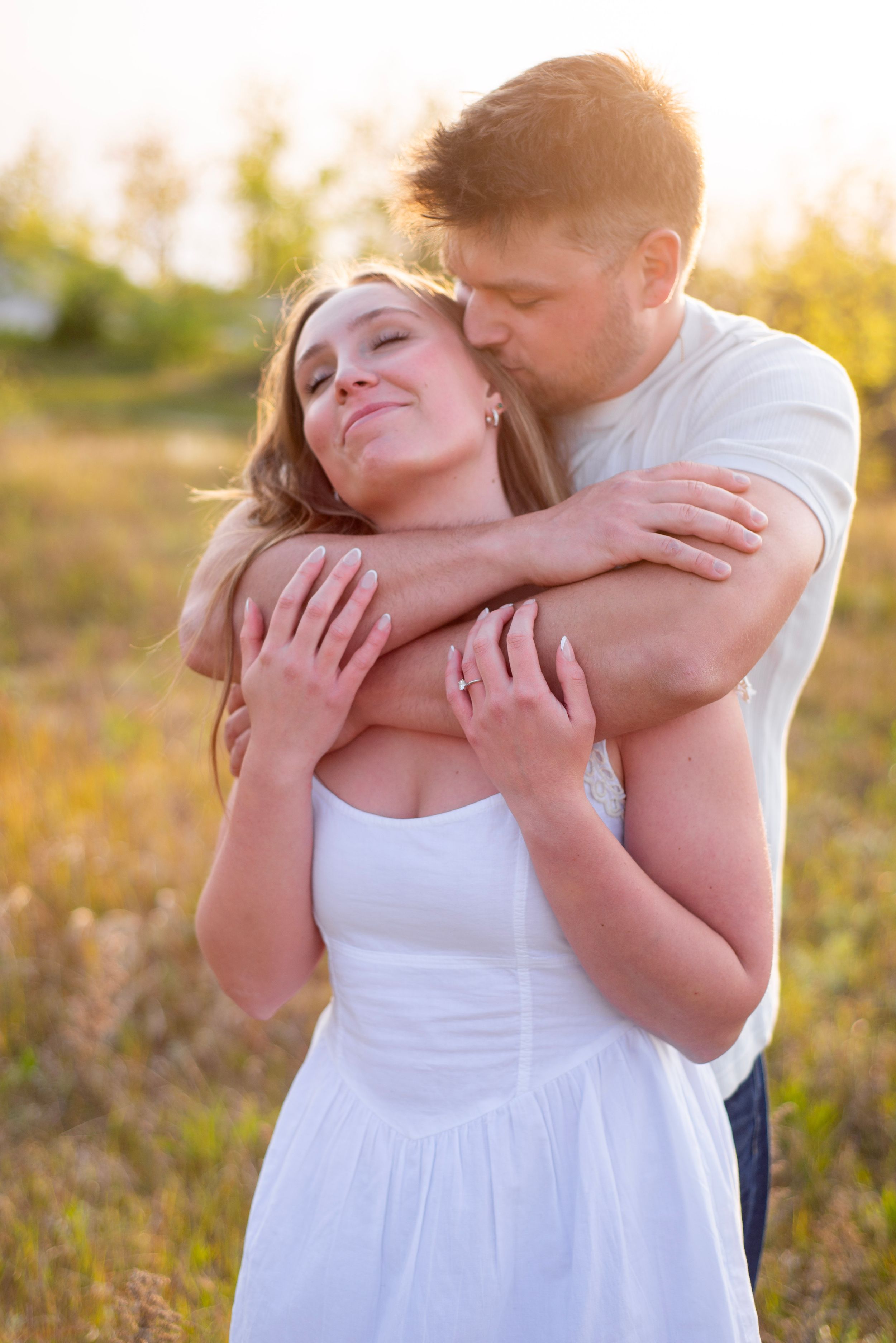 A couple embraces in a sunlit field, man kissing woman's cheek as she smiles in a white dress.