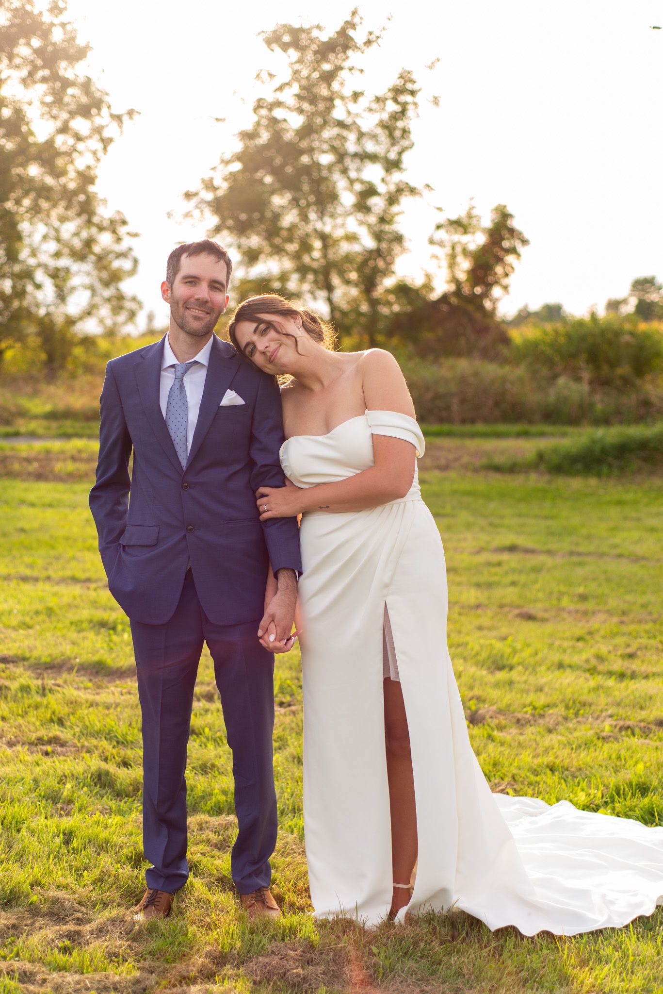 A couple poses together in an outdoor field at sunset, with her in a white gown and him in a navy suit.