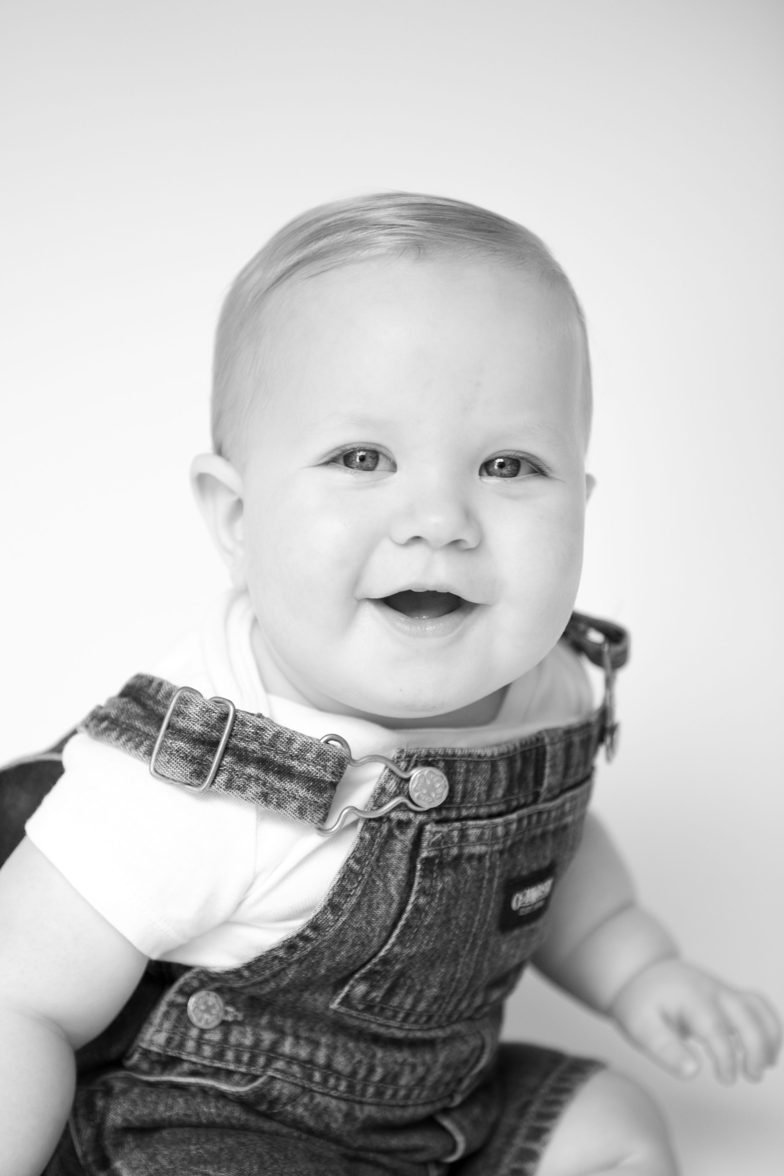 Baby wearing denim overalls photographed in black and white against white backdrop.