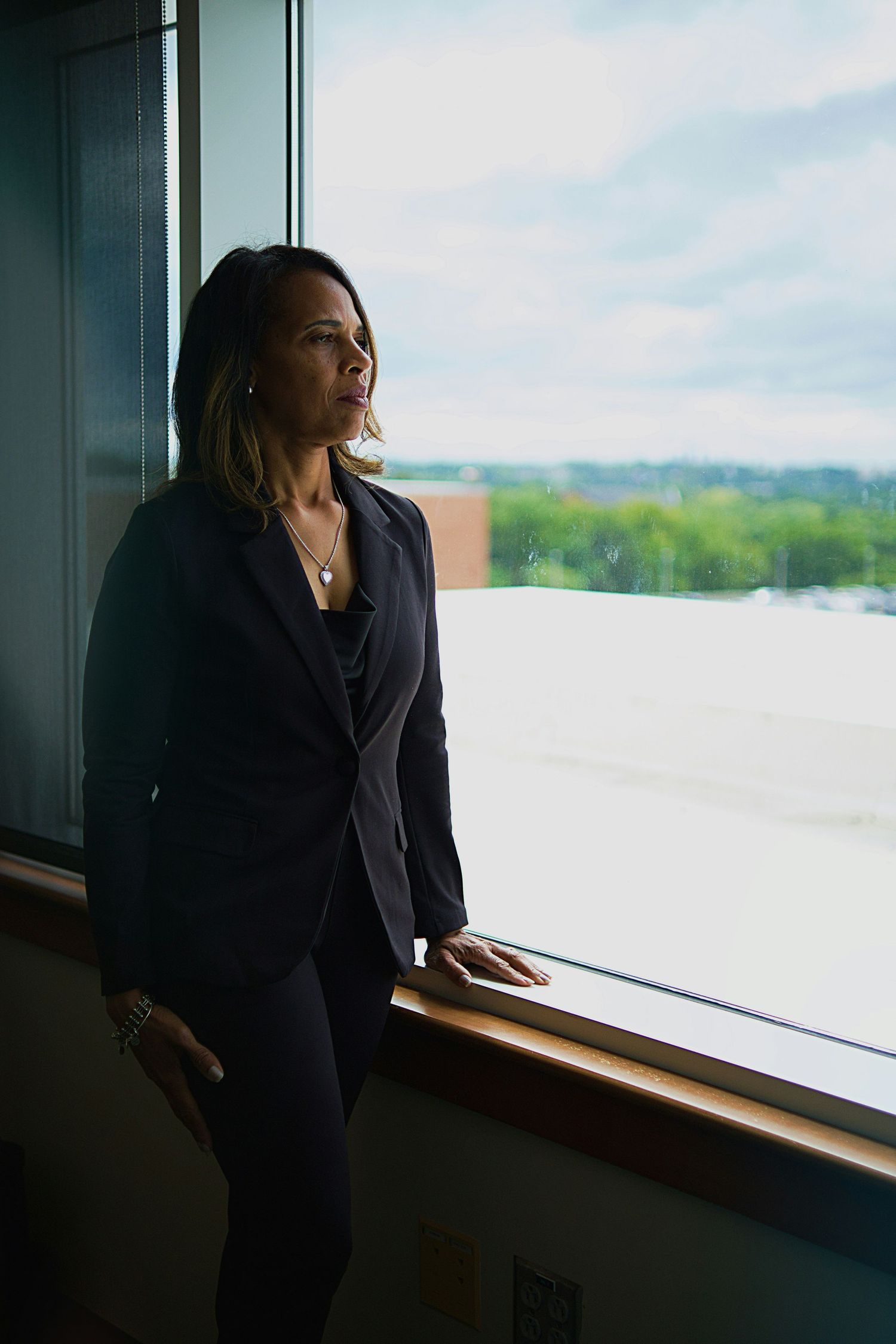 Professional in dark blazer gazing thoughtfully through office window at city view.